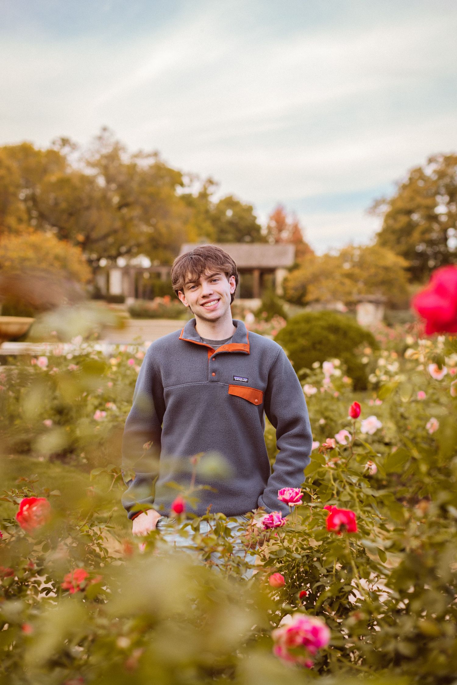 A young man in a blue pullover smiles for the camera surrounded by roses during his senior photo session in Kansas City, Missouri.