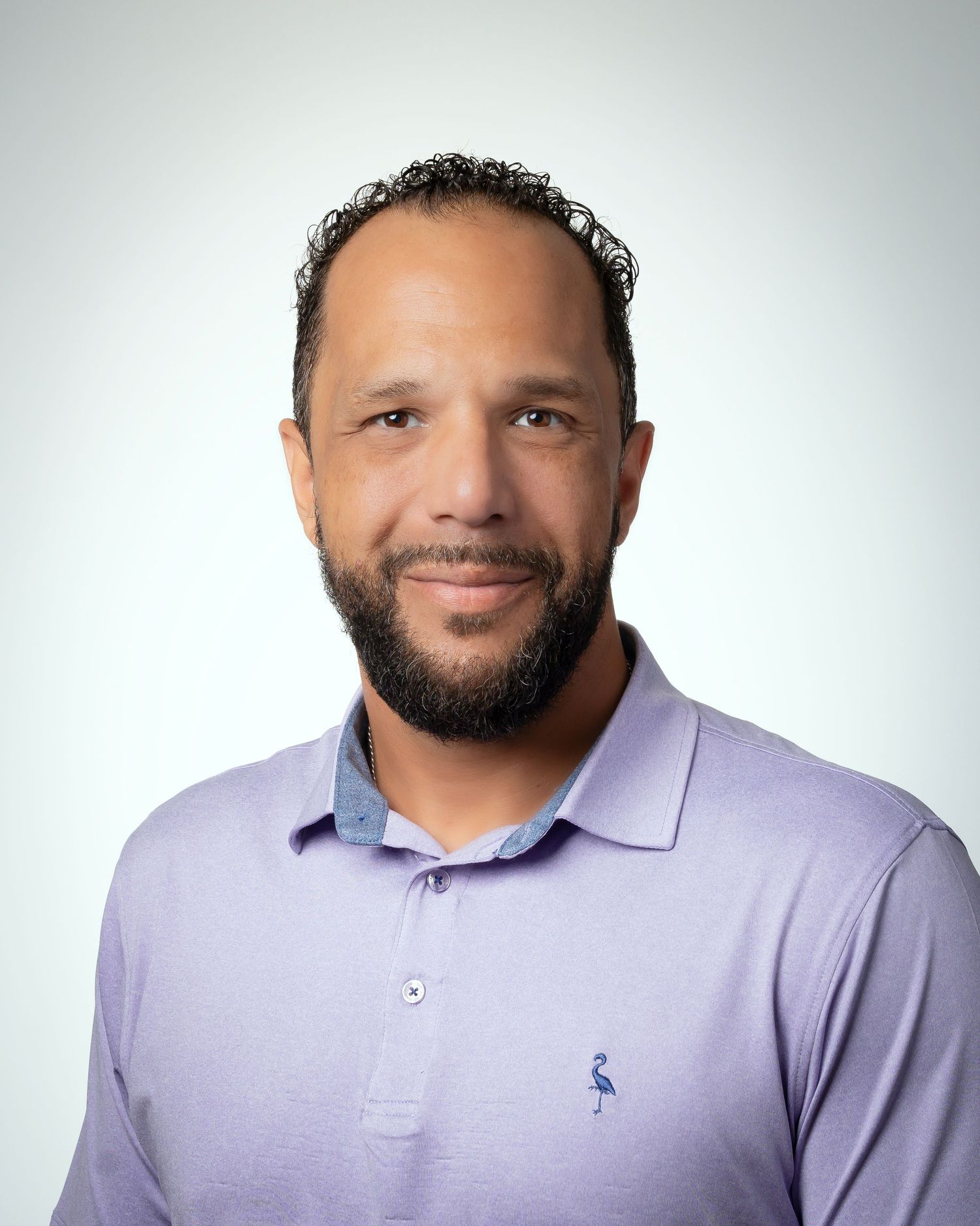 A person, Garrett Maney, in a self-portrait, wearing a lavender polo shirt, smiles warmly against a light gray studio background.
