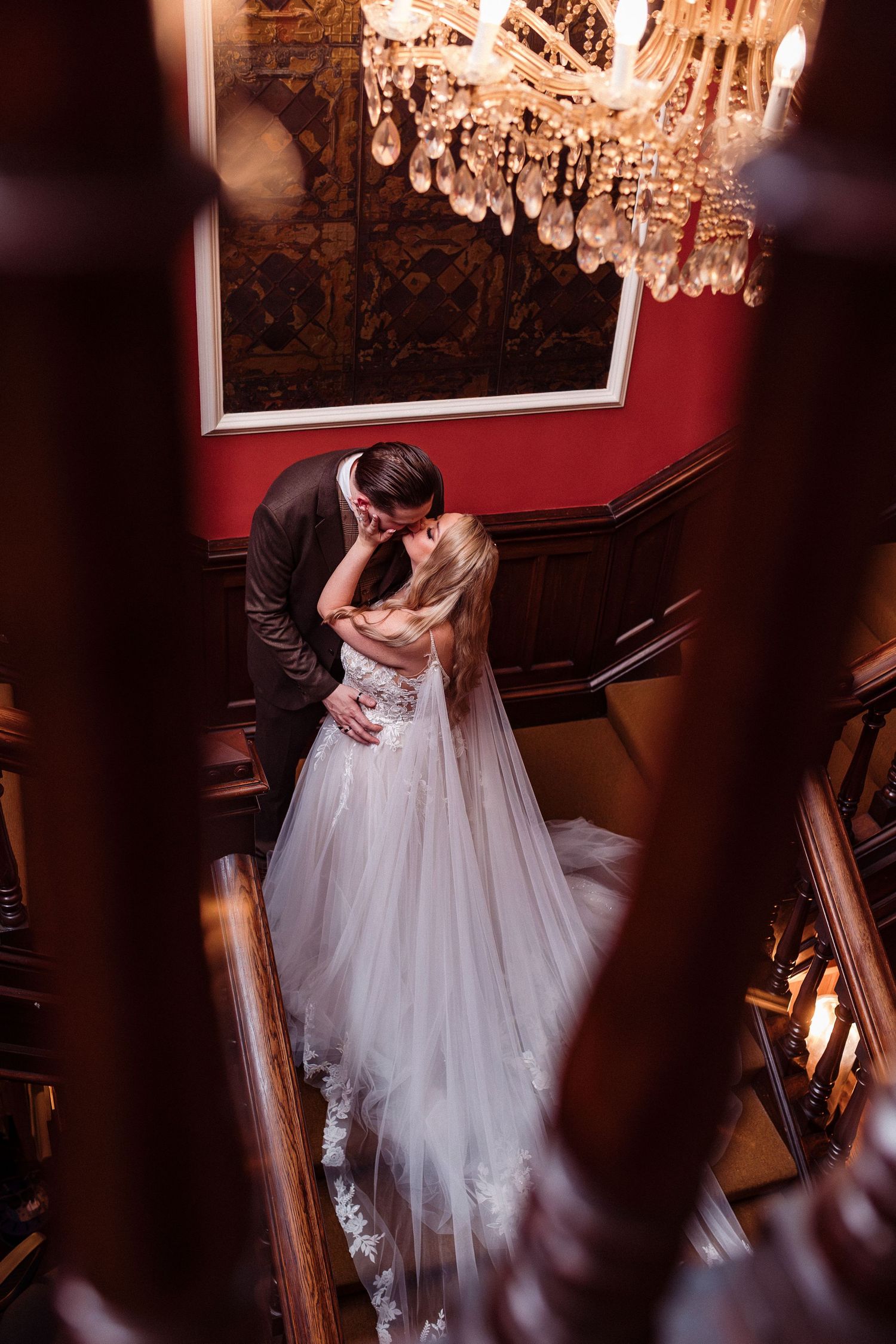 A romantic couple embraces under a crystal chandelier in a dimly lit room with red walls and vintage decor.