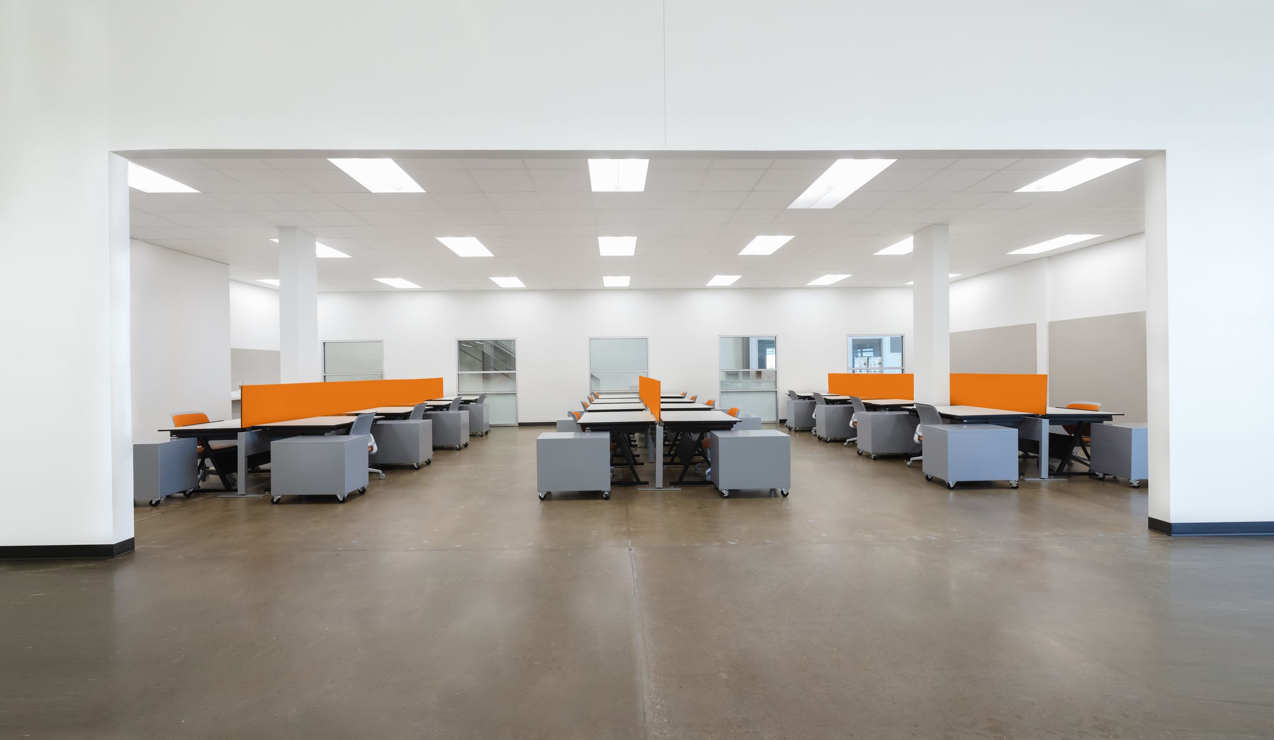 Large empty office space with white walls, fluorescent lighting, and orange accents on workstations.