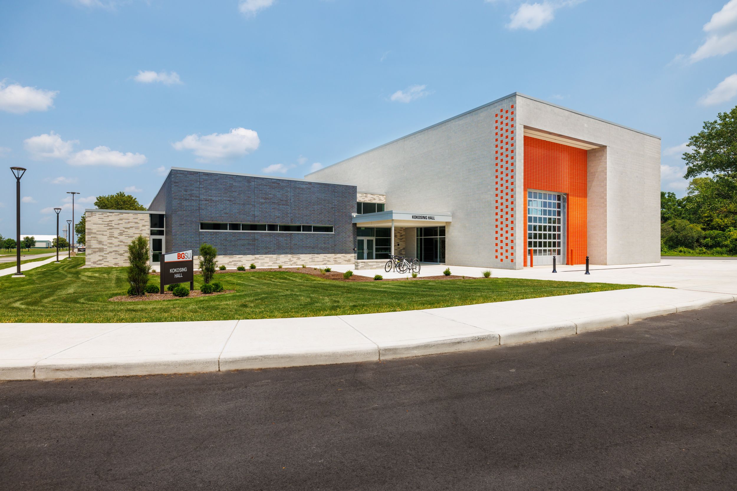 Modern industrial building exterior with orange accents and curved driveway against blue sky and green landscaping.
