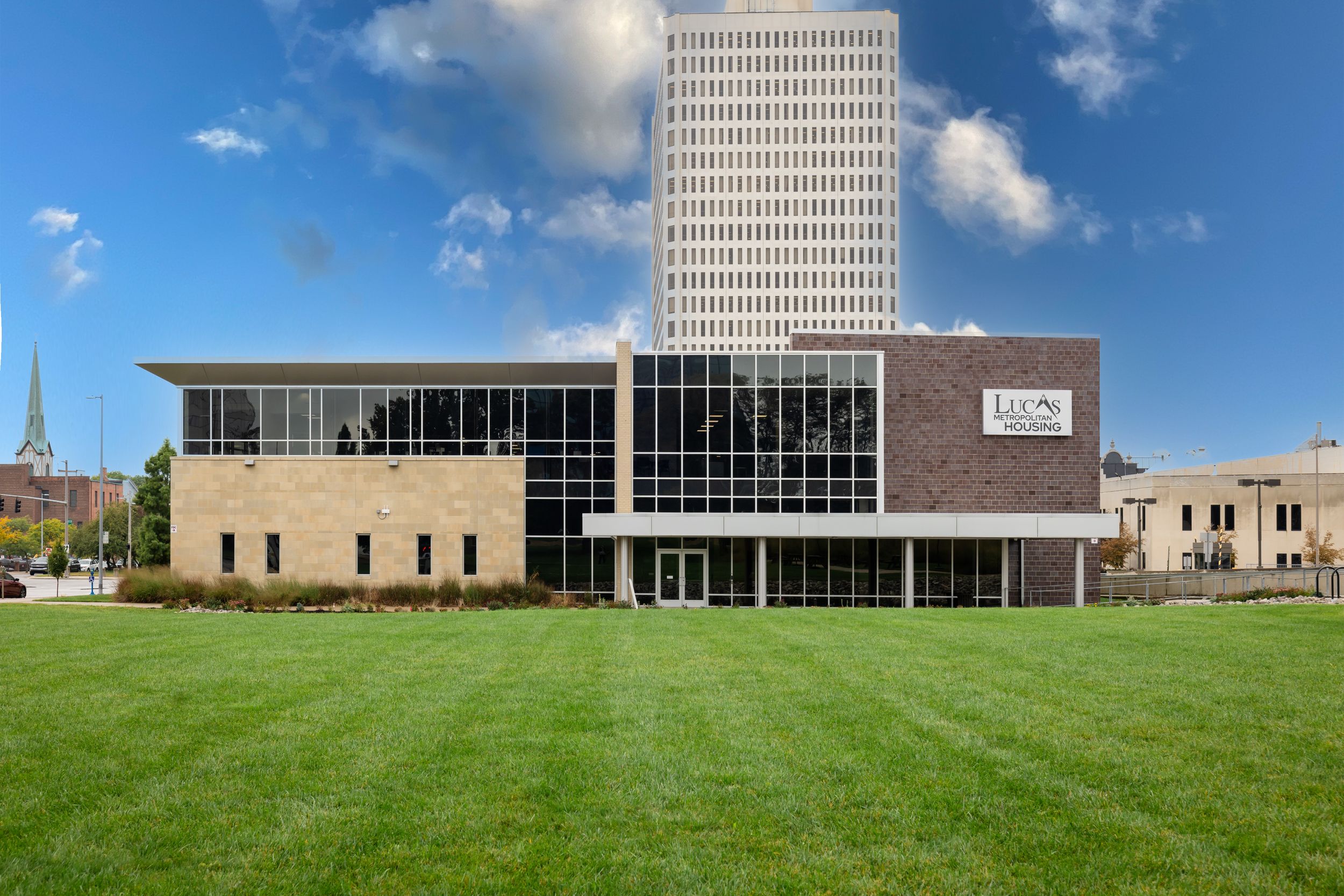 Modern office building with glass facade and tall tower rises behind expansive green lawn on sunny day.