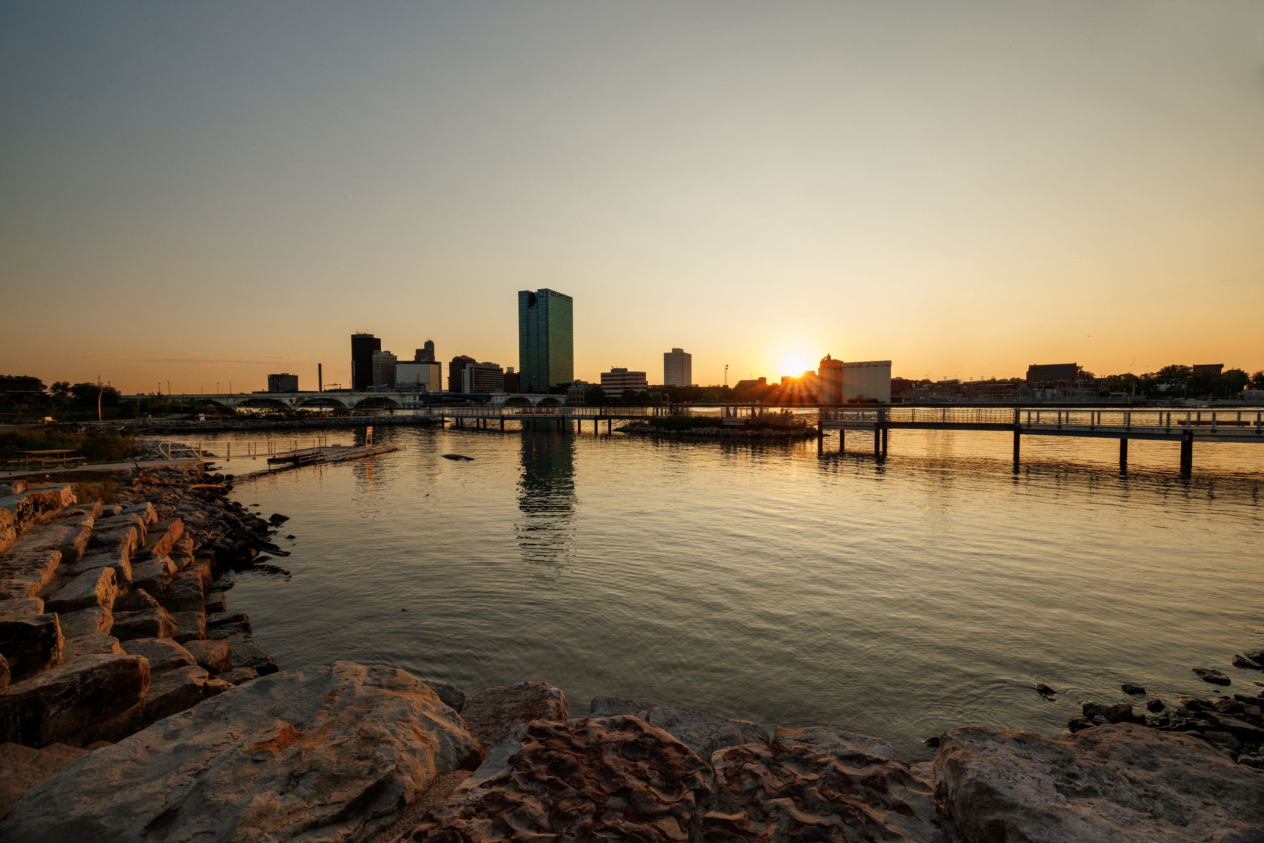 Panoramic view of a city skyline at sunset reflecting on calm harbor waters.