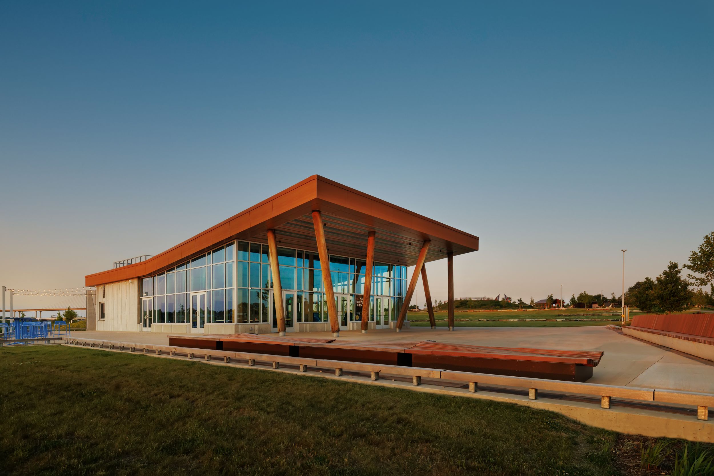 Modern glass pavilion with angled wooden roof and curved benches at sunset against clear blue sky.
