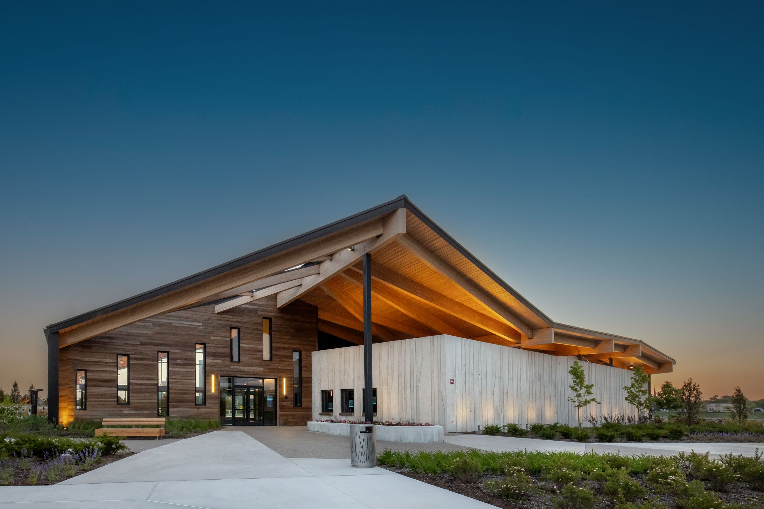Modern architectural building with angular roof lines and glass walls illuminated at sunset against a blue sky.