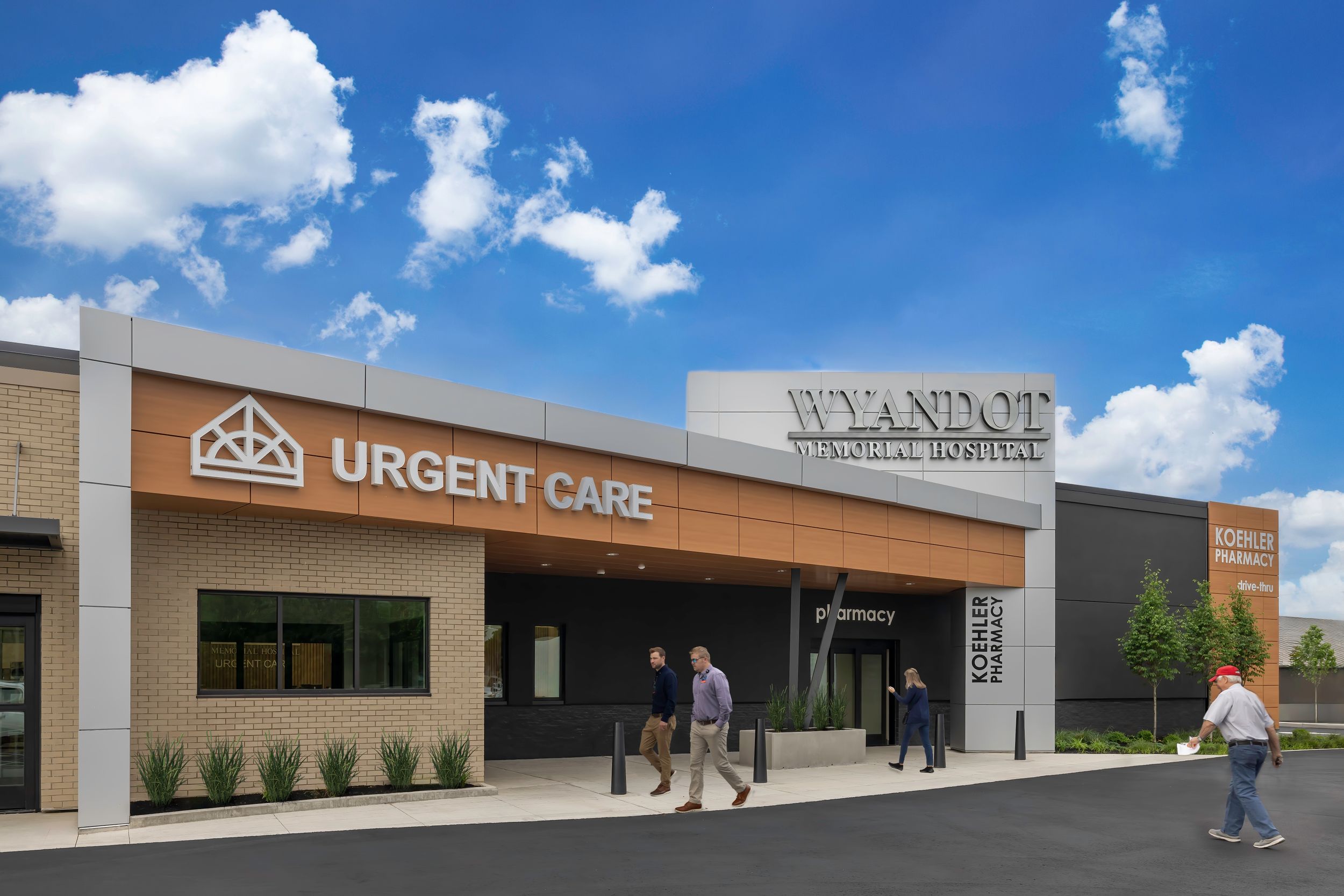 Exterior view of an urgent care medical facility in a strip mall with people walking outside under blue skies.