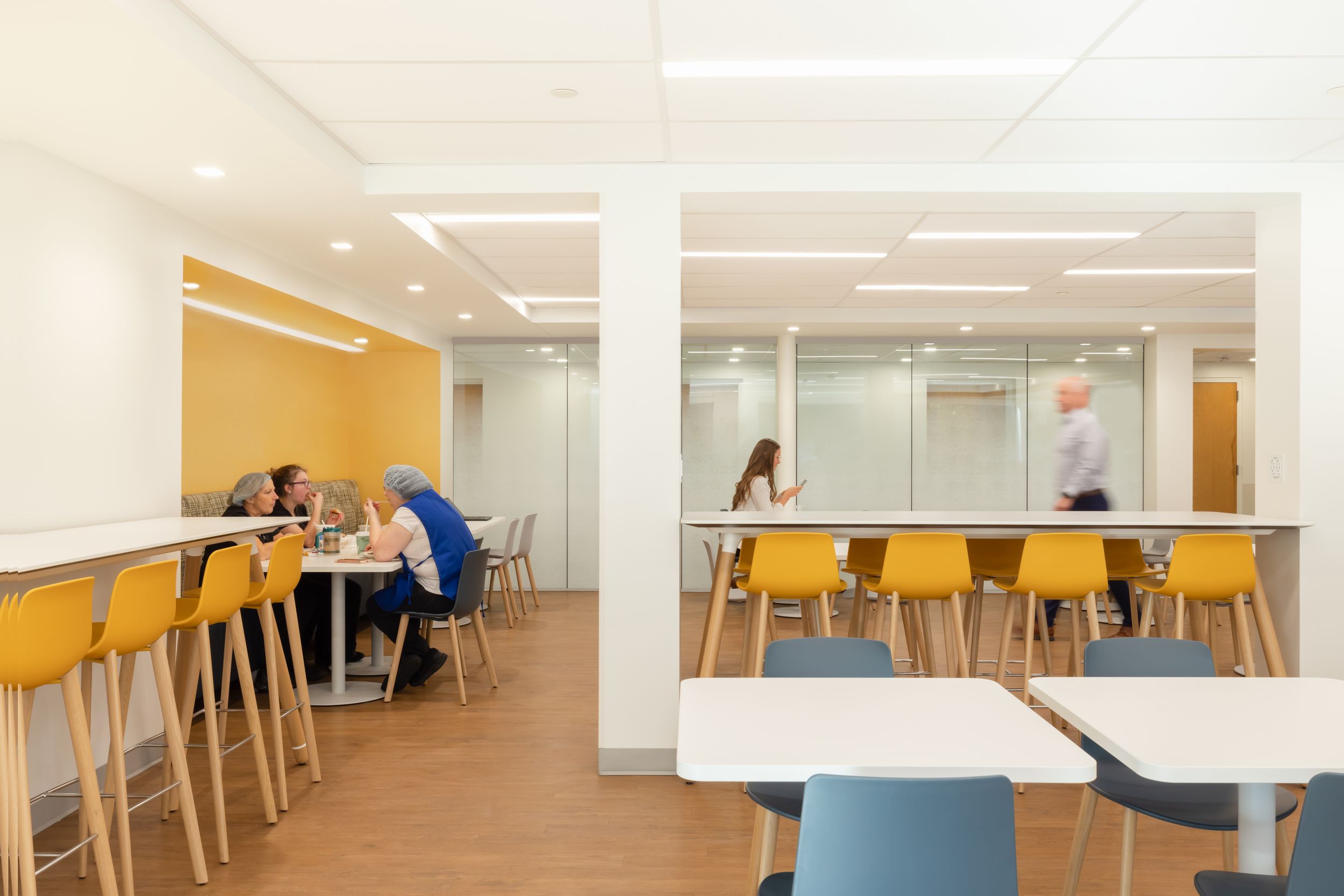Modern cafeteria space with wooden tables and chairs features bright lighting and yellow accent walls.