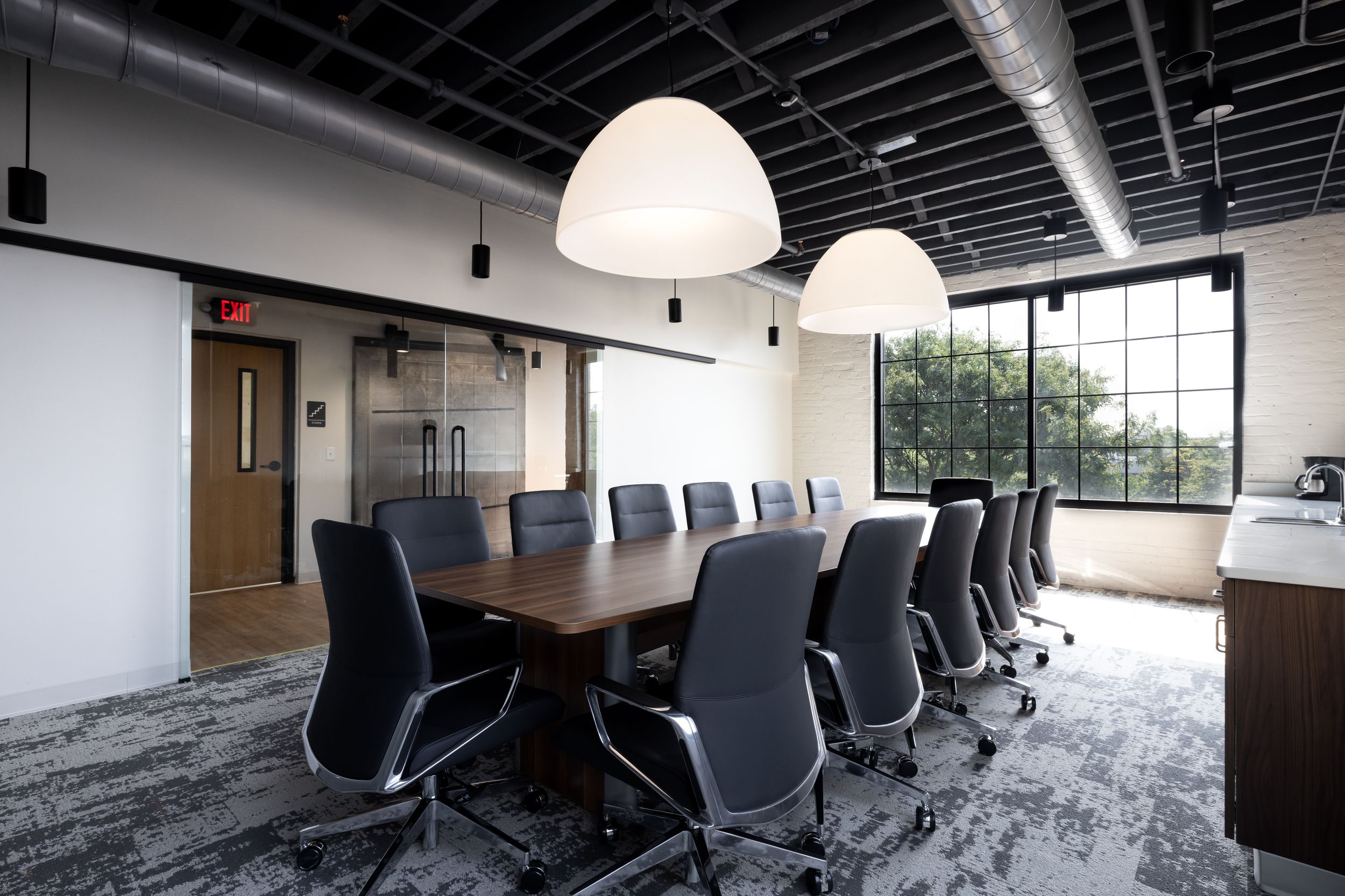 Modern conference room with long wooden table, black office chairs, white pendant lights and exposed ceiling ductwork.