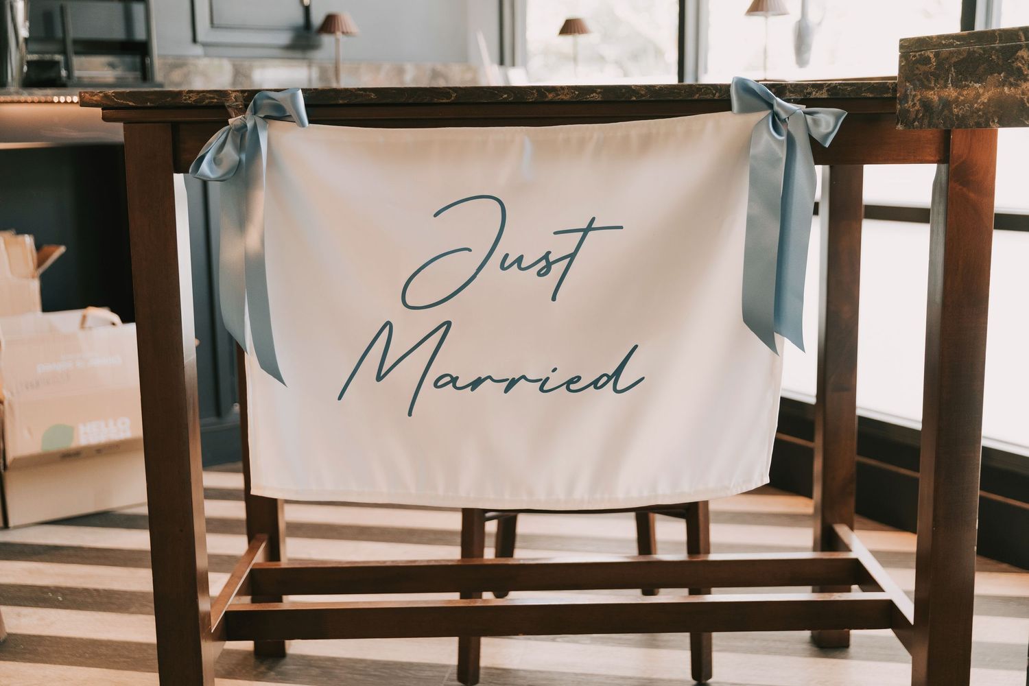 White fabric chair banner with 'Just Married' text and blue ribbons hangs on wooden chair at wedding reception.