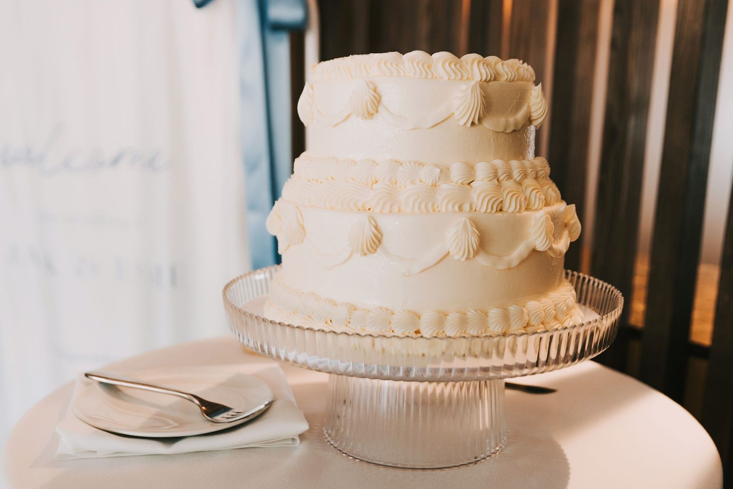 A classic three-tiered white wedding cake with delicate ruffled edges sits on a glass pedestal cake stand.