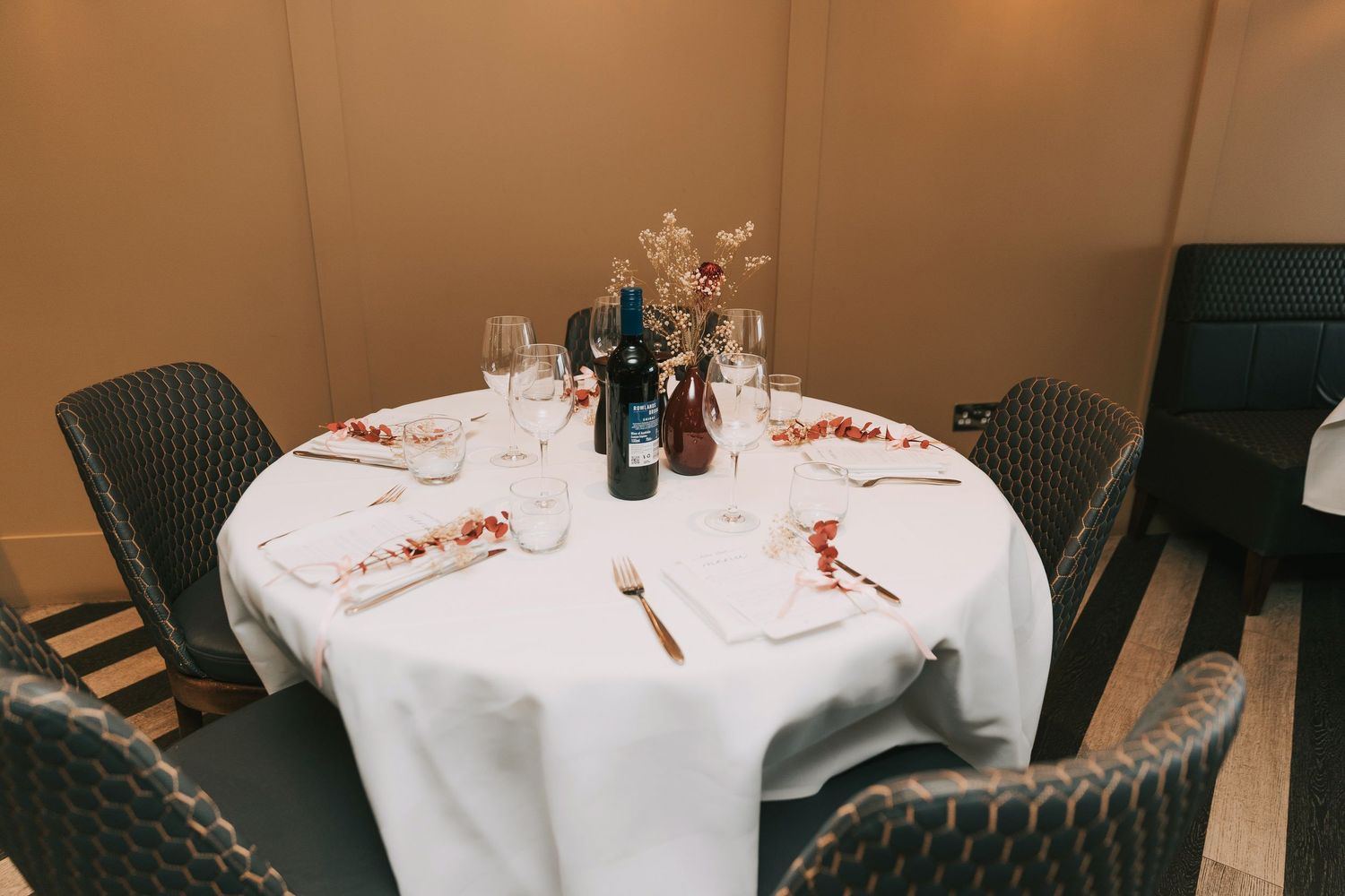 A round dining table set with white linens, wine glasses, and wicker chairs in an intimate restaurant setting.