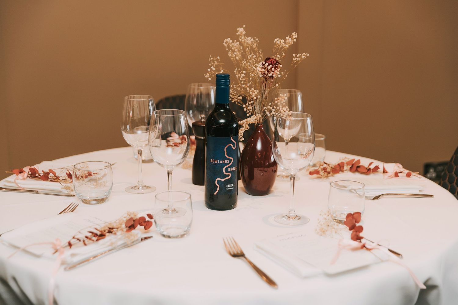 Elegant table setting with wine bottle, glasses, and dried floral centerpiece on white tablecloth.