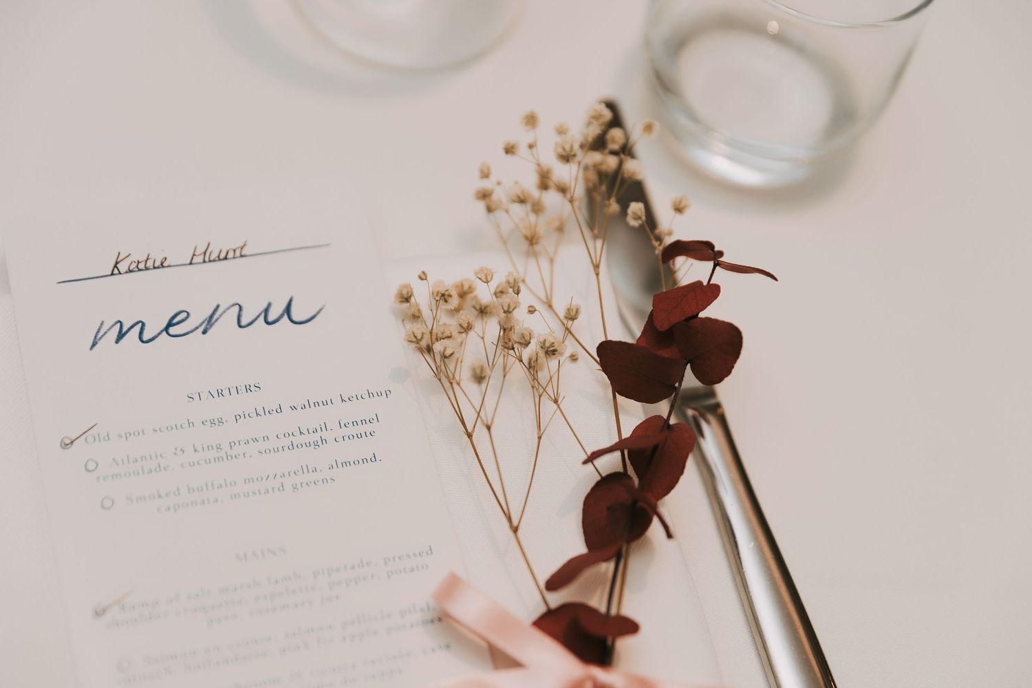 Elegant wedding menu cards and floral decor arranged on a soft pink surface with dried flowers and delicate ribbons.