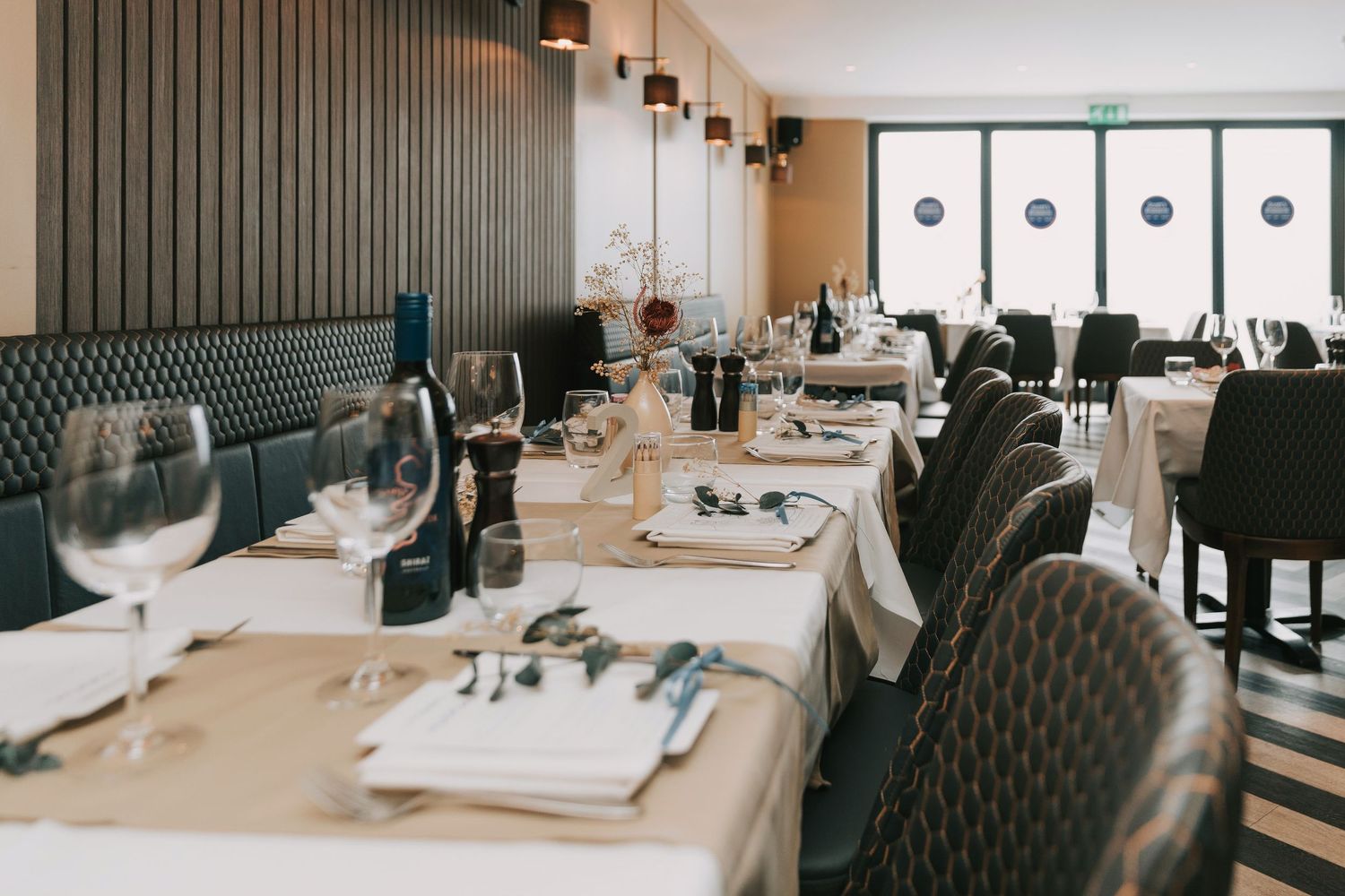 Modern restaurant interior with white tablecloths, tufted gray chairs, and large windows offering natural light.
