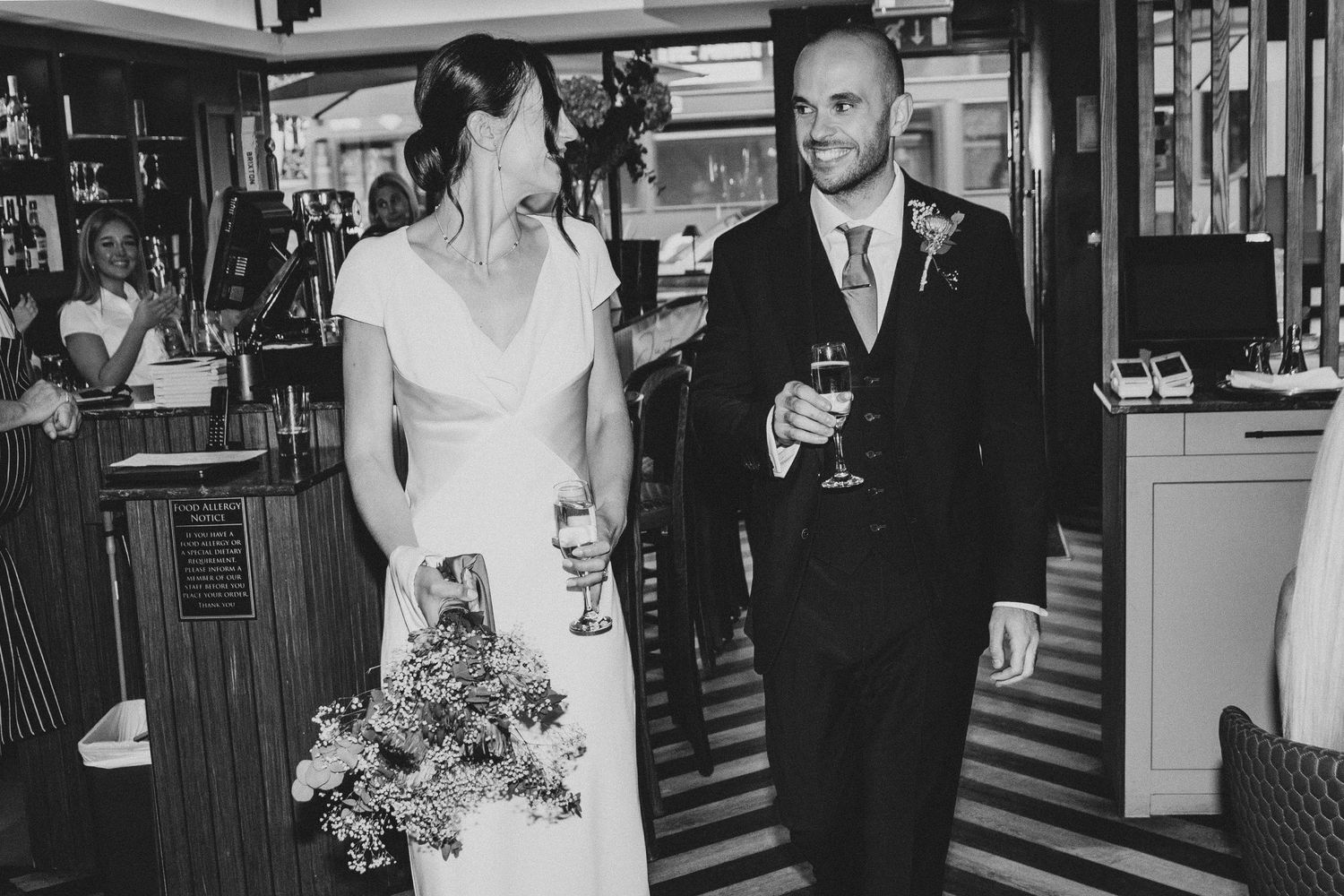 A bride and groom share drinks together at a vintage bar in a black and white photograph.
