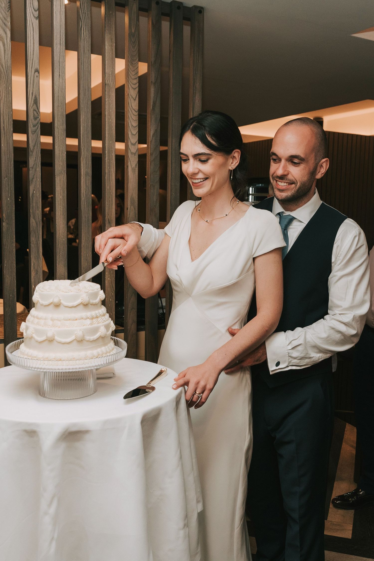 Wedding couple and guests share joyful moments while cutting and serving a white tiered wedding cake.