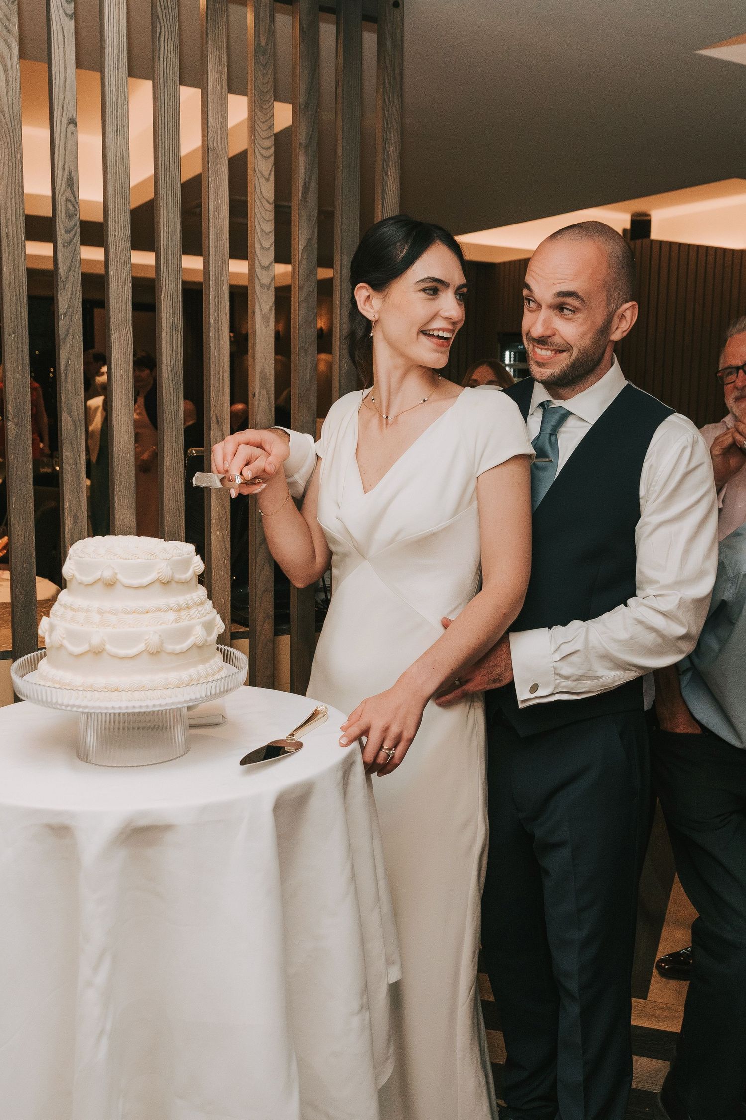 A wedding couple shares a playful moment while cutting their elegant white tiered cake at their reception.
