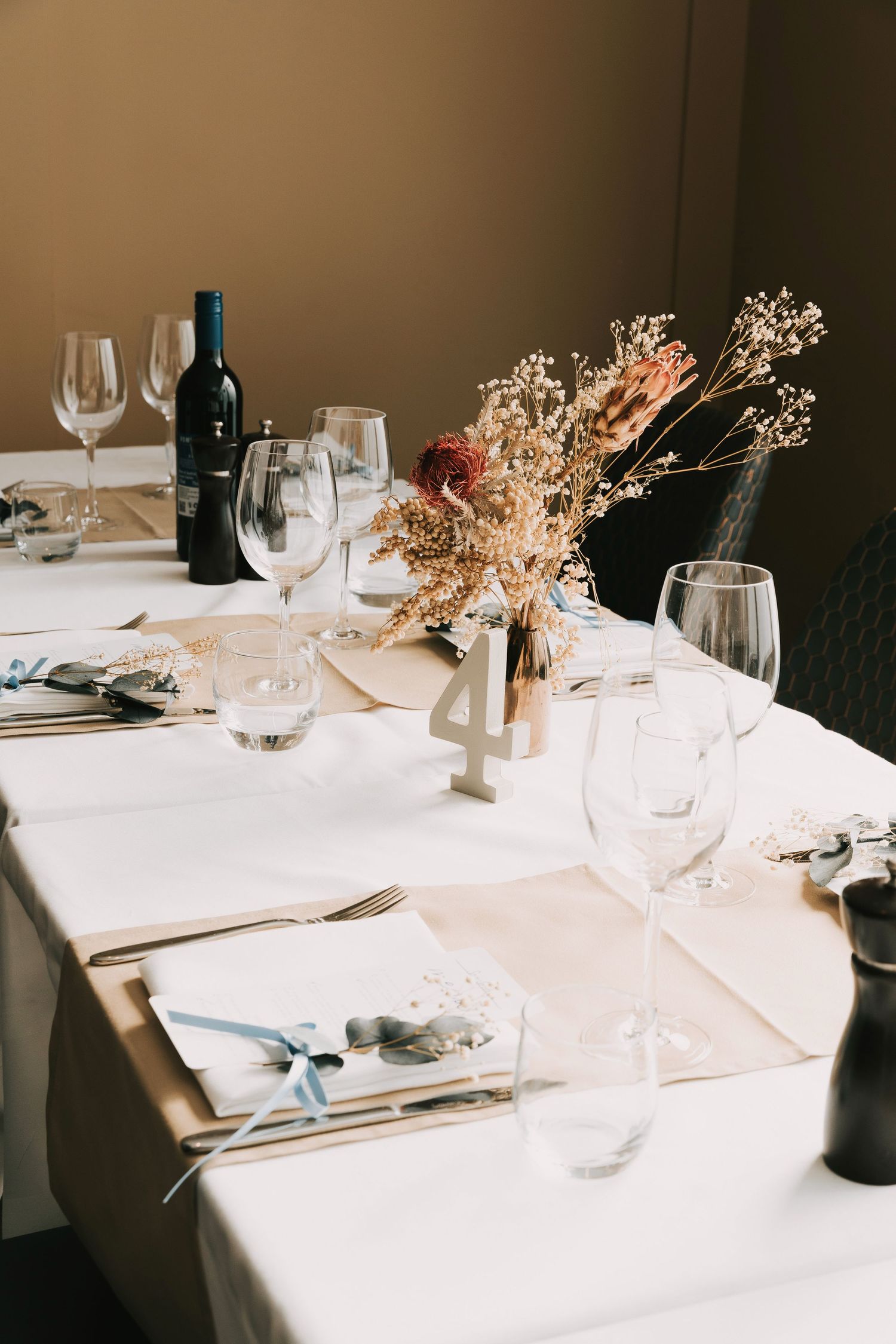 Elegant table setting with white linens, wine glasses, and dried floral centerpiece in warm lighting.