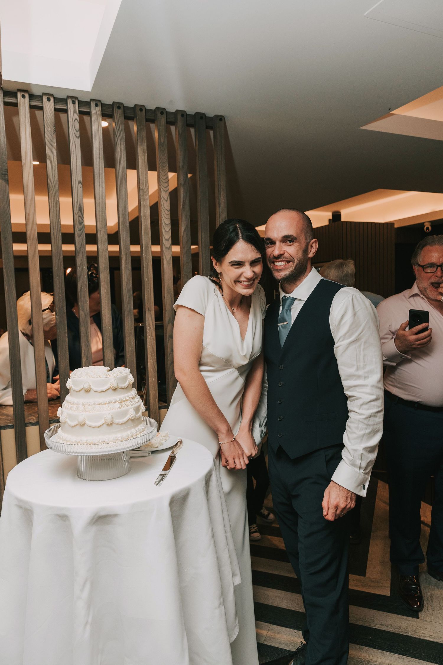 Wedding celebration scene showing guests gathered around a white tiered cake in an elegant indoor venue.