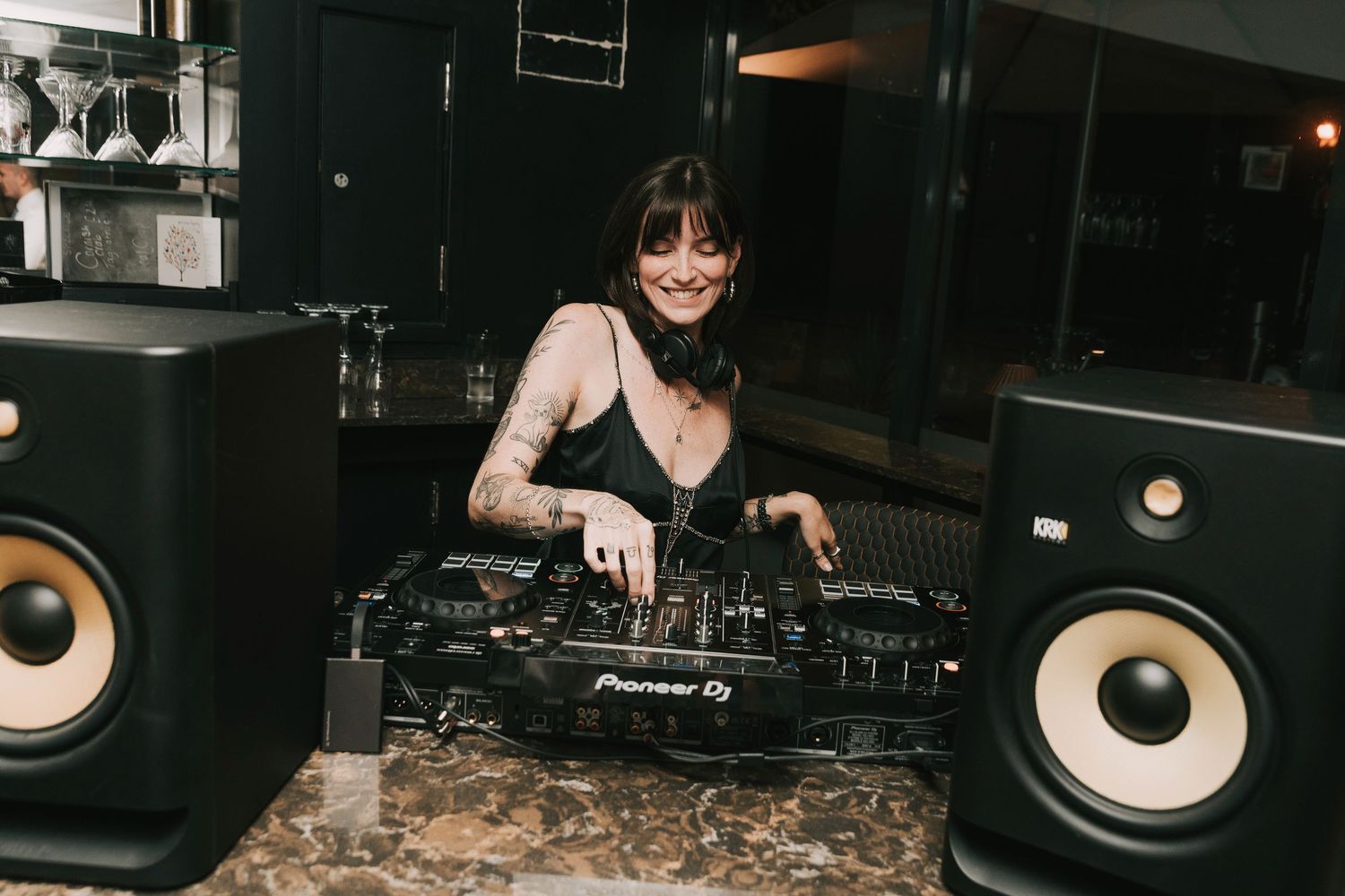A DJ performs at a dark nightclub setting with Pioneer equipment and large speakers visible.