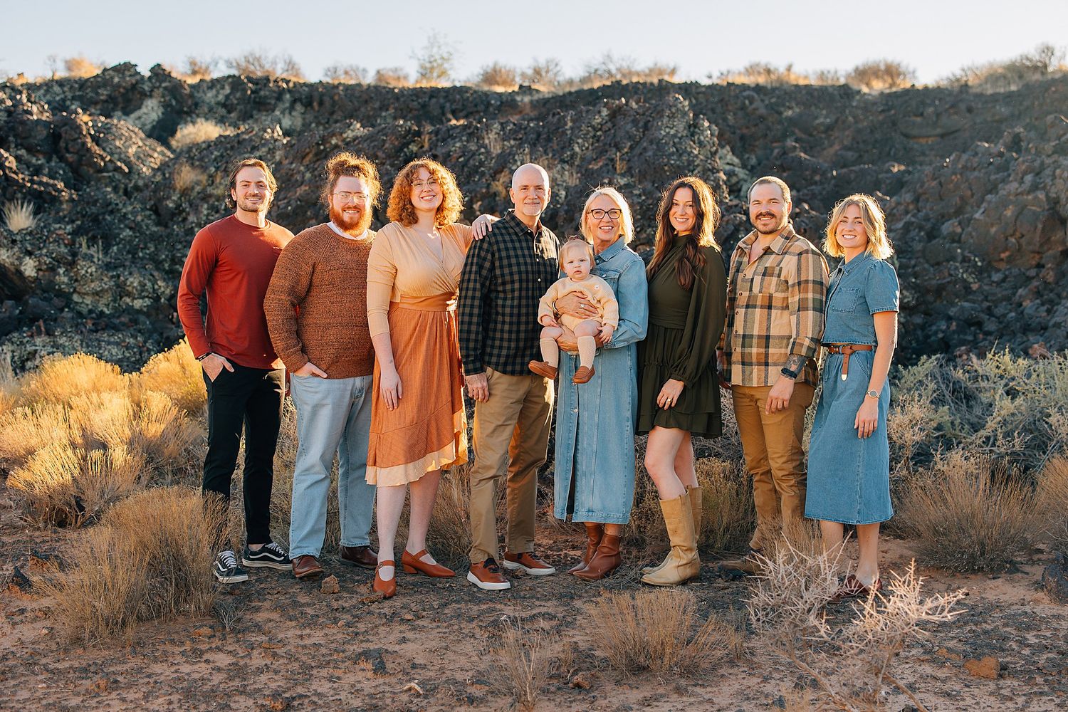 A group of people wearing earth-toned clothing stand together in front of a desert landscape at sunset.