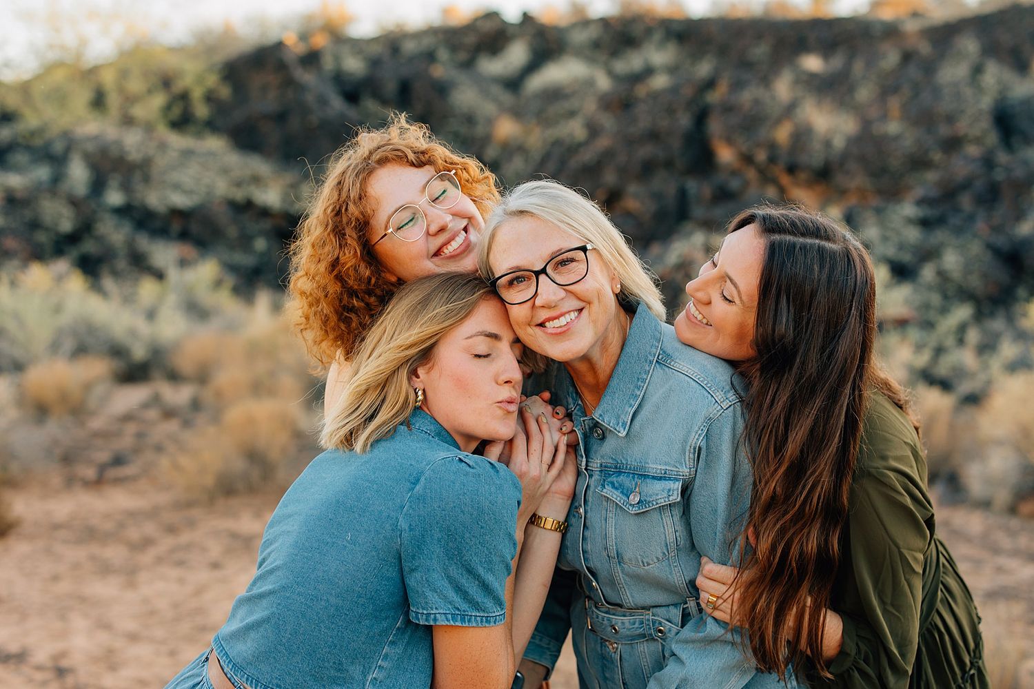 Three people laughing and embracing while posing outdoors in casual clothing against a desert landscape background.