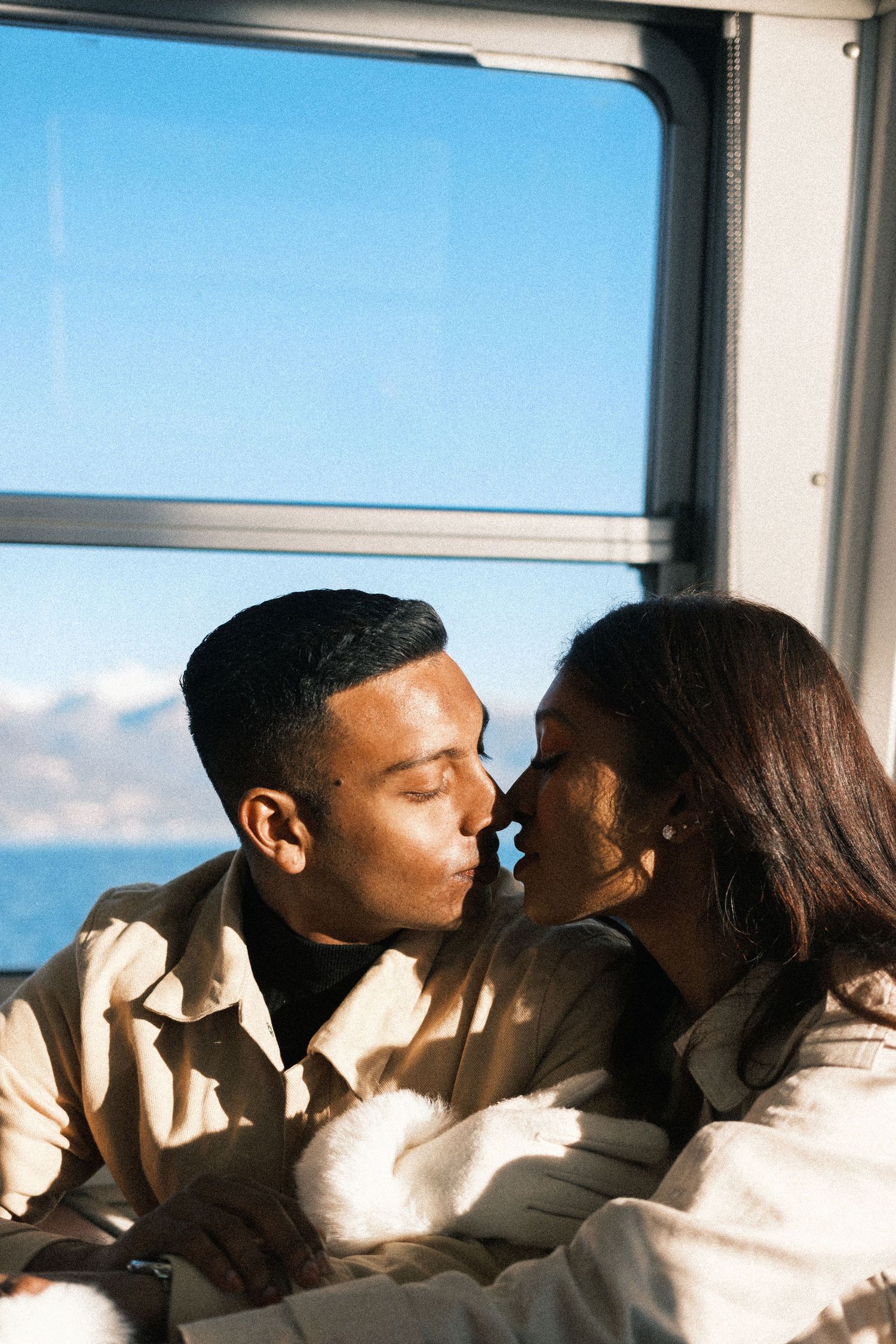 A couple shares a tender kiss while sitting by a window with bright blue sky visible in the background.