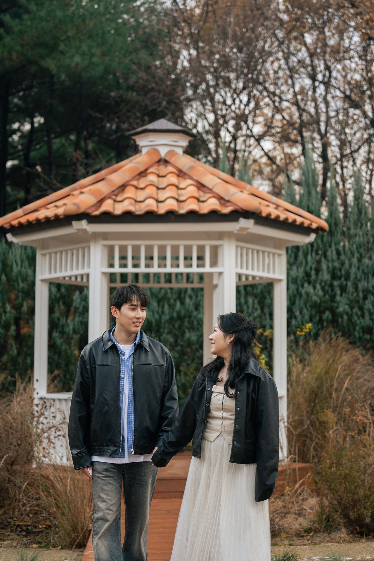 A couple stands together in front of white gazebos and autumn trees in a romantic outdoor setting.
