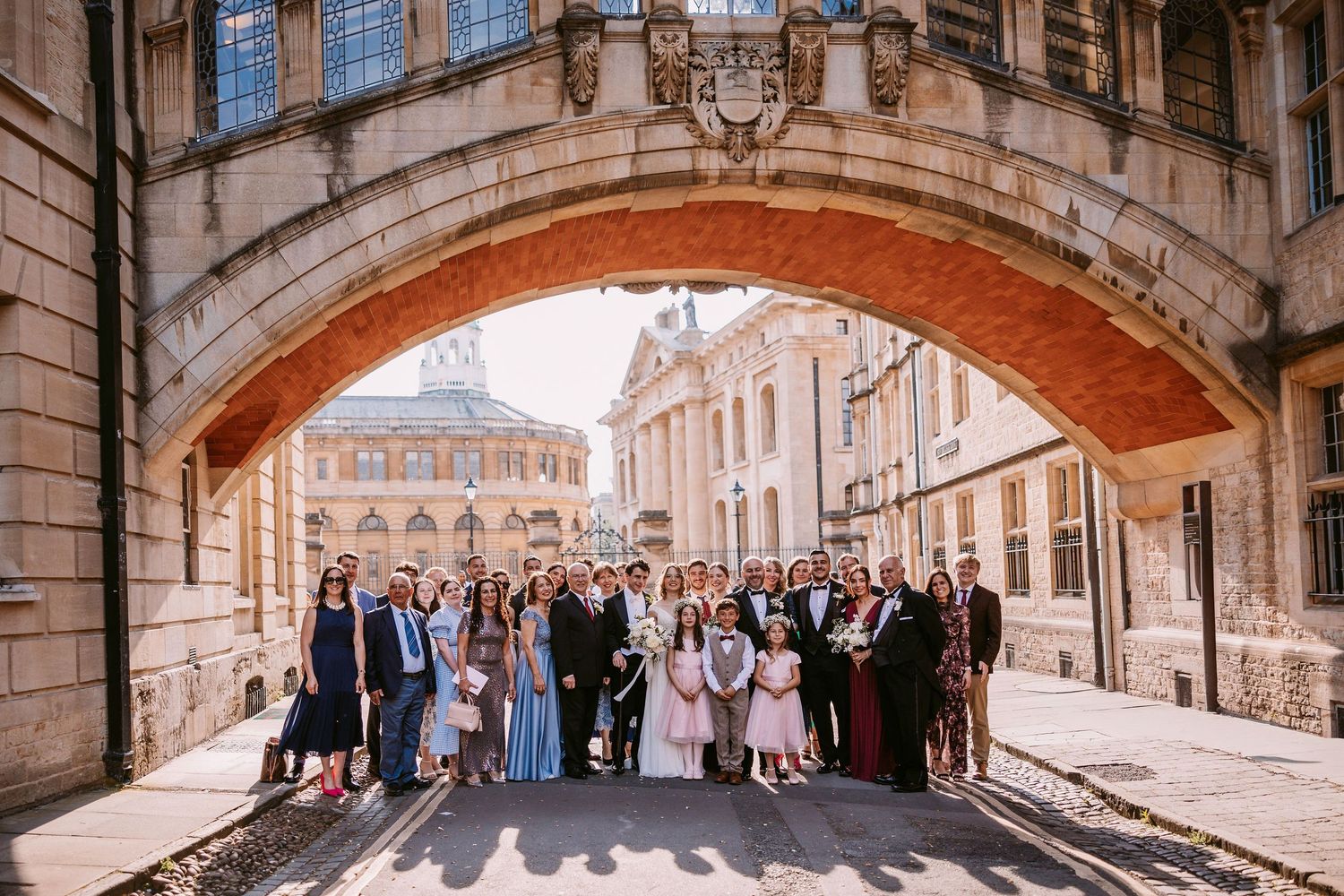A group of formally dressed people pose under a historic stone archway in Oxford, England.