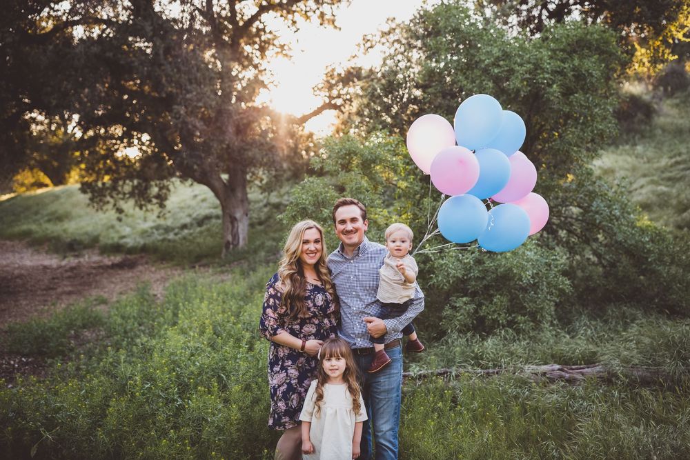 Family holding pink and blue balloons poses together in a sunlit outdoor garden setting during golden hour.