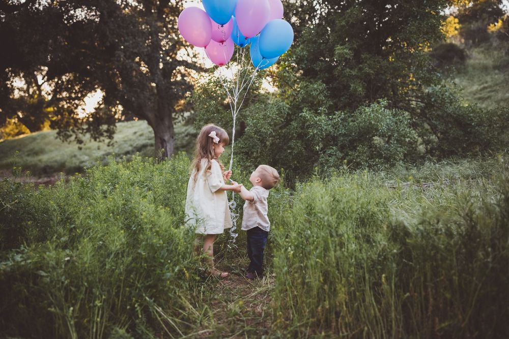 Two people stand in tall grass at sunset holding pink and blue balloons in a dreamy outdoor setting.