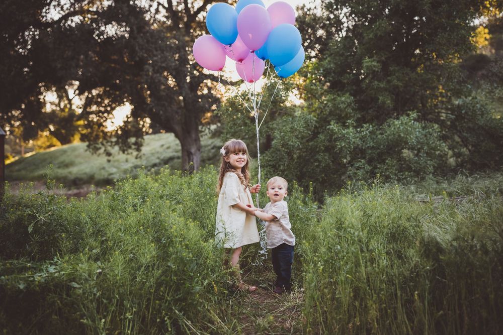 A colorful outdoor photoshoot scene with pink and blue balloons floating among trees in a natural setting.