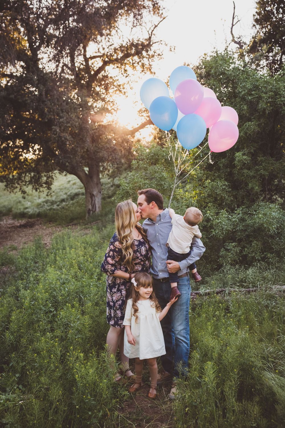 Family holding blue and pink balloons poses together in a natural outdoor setting during golden hour sunset.