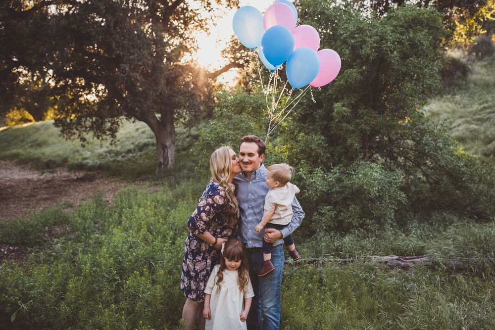 Family holds pink and blue balloons during a sunset photoshoot in a wooded outdoor setting.