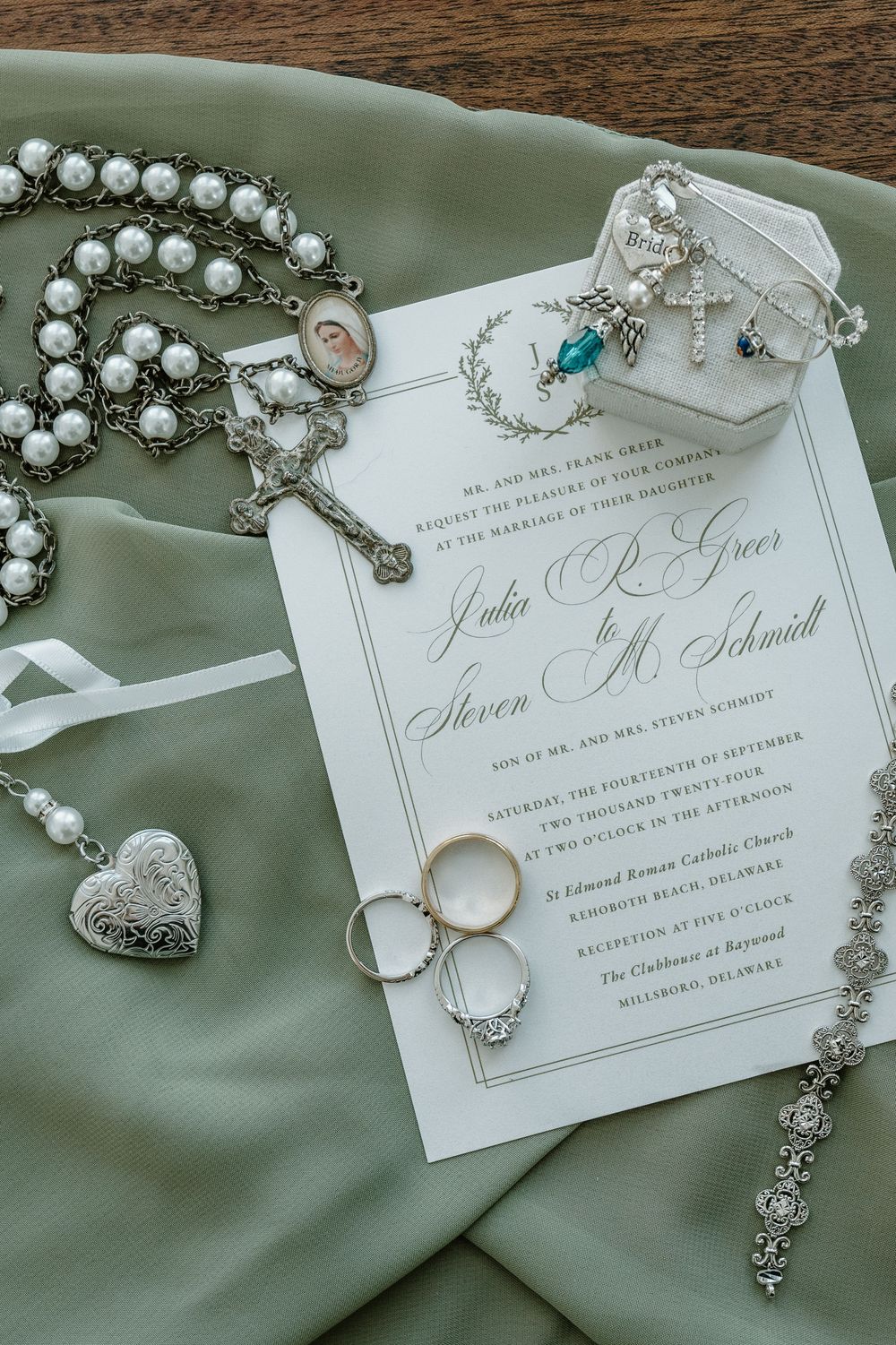Elegant wedding invitation with pearl necklace and diamond rings on sage green fabric.