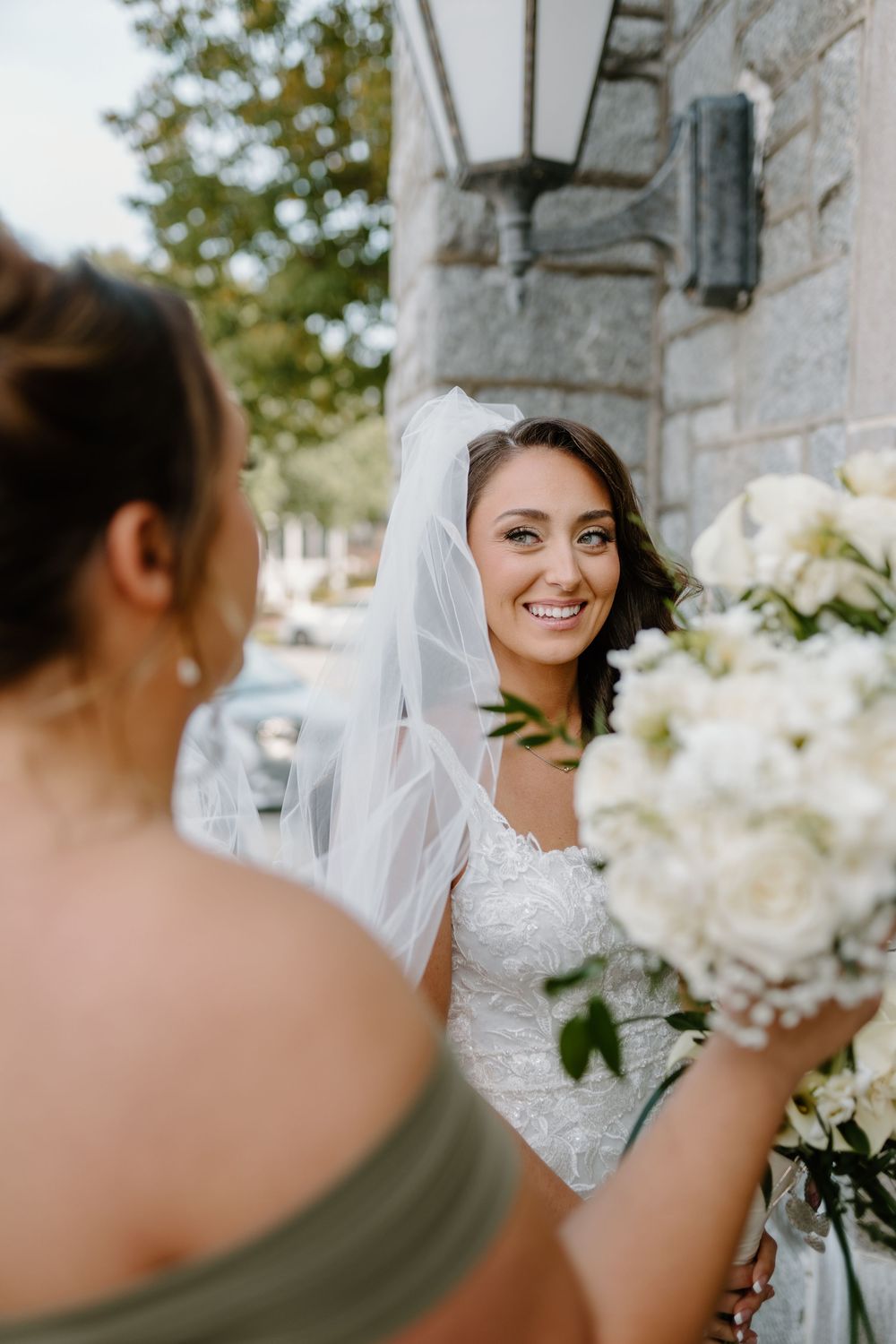 A bride holding a white floral bouquet stands outside a stone building.