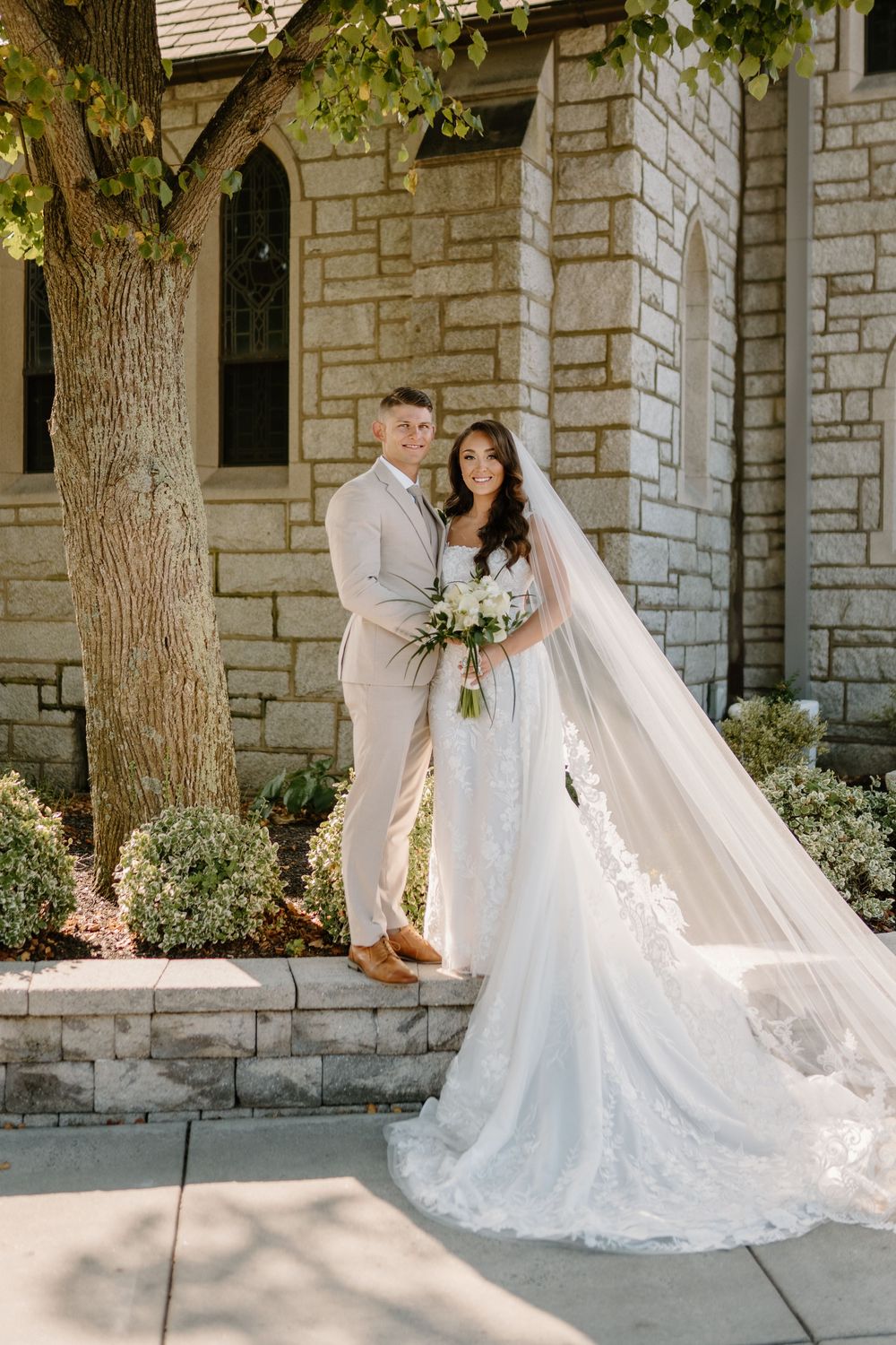 Elegant bride in white lace gown with flowing veil poses against stone church exterior.