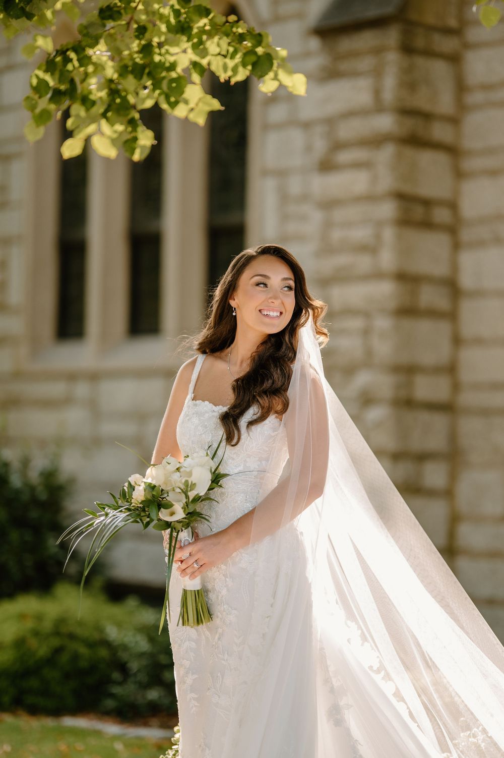 A flowing white wedding dress and veil catch the sunlight against a stone building.