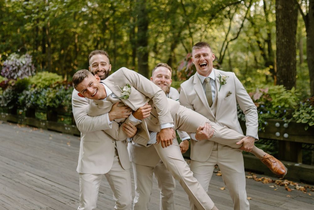 Groomsmen in light beige suits playfully pose together on a wooden deck in a garden setting.