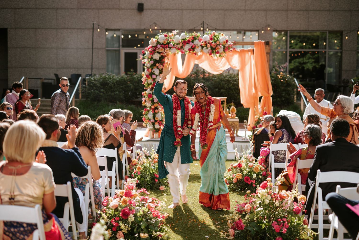 Wedding guests celebrate at an outdoor garden ceremony decorated with pink flowers and coral drapes.