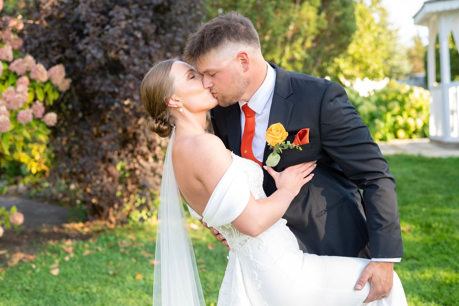 A romantic wedding couple shares a kiss in a lush garden setting during their summer celebration.
