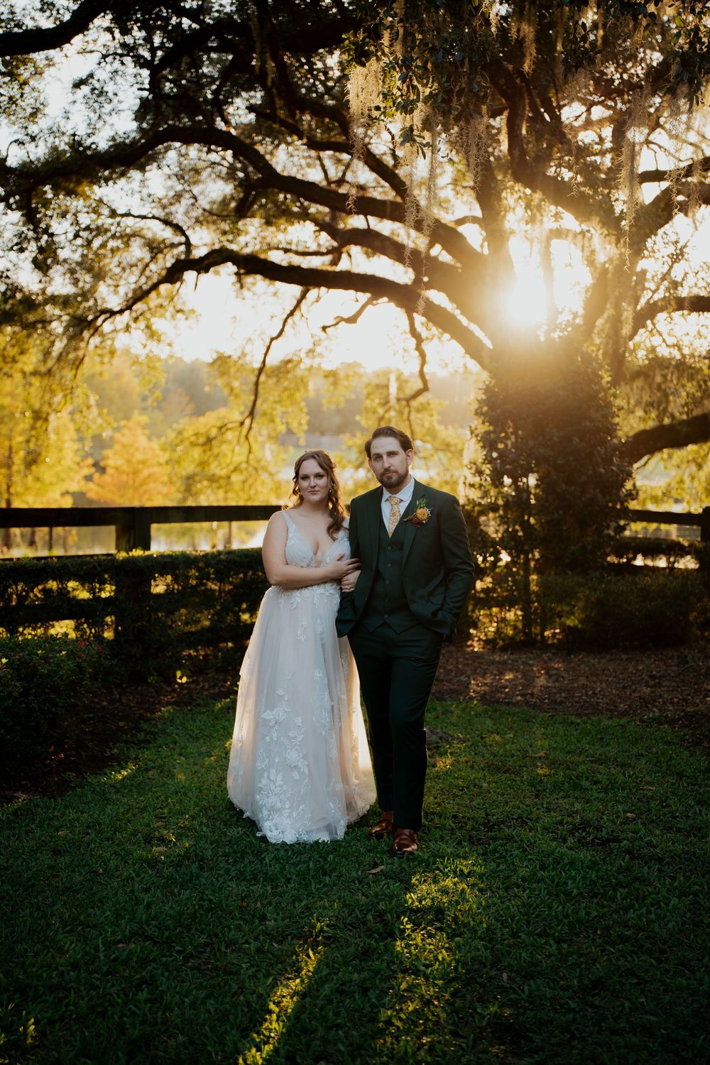 A couple poses together at sunset under a majestic oak tree with Spanish moss, backlit by golden sunlight.