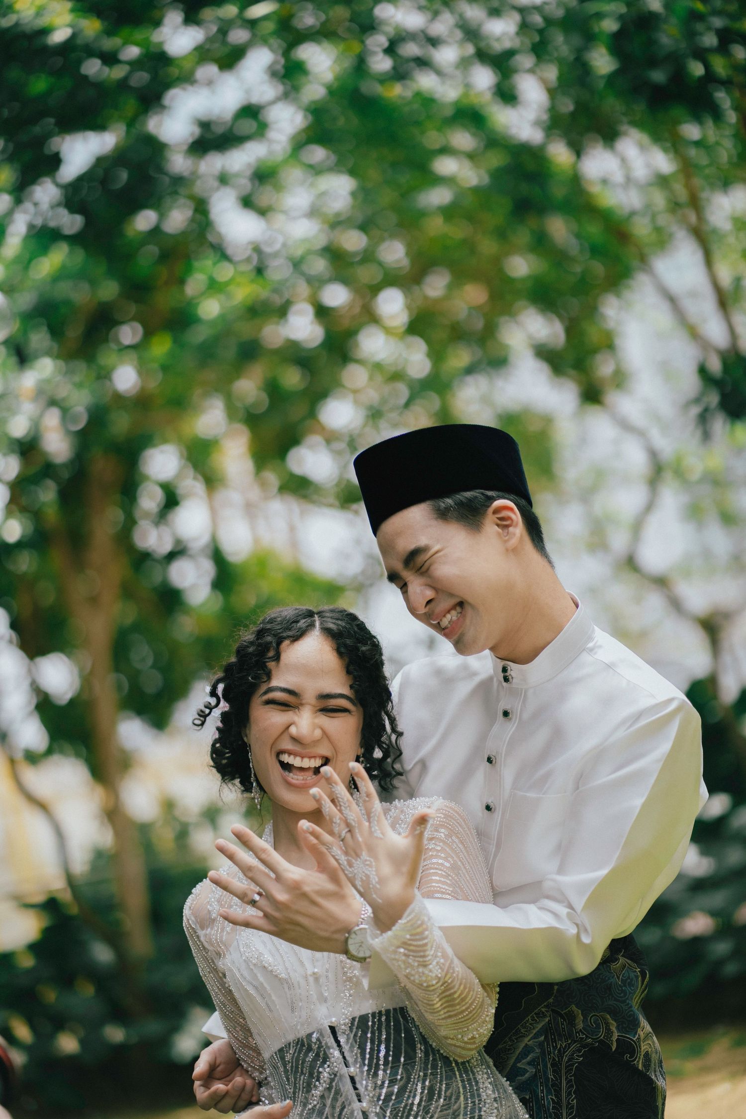 Laughing Malay couple in traditional attire proudly display wedding rings outdoors.