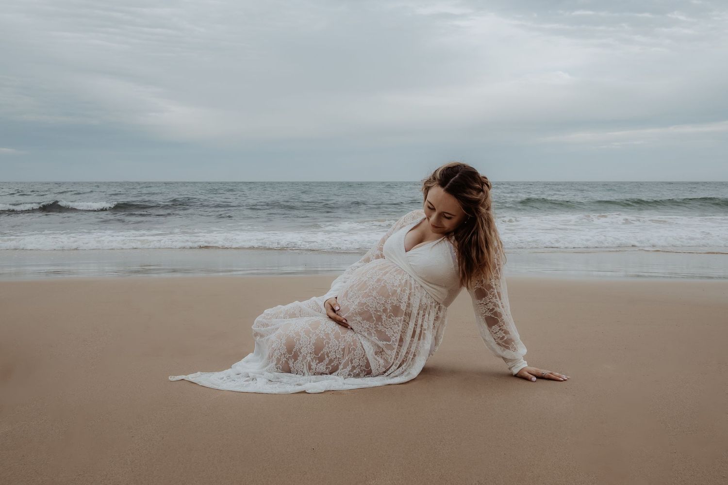 Pregnant woman in white dress posing gracefully on a sandy beach at sunset during a central coast maternity photoshoot.
