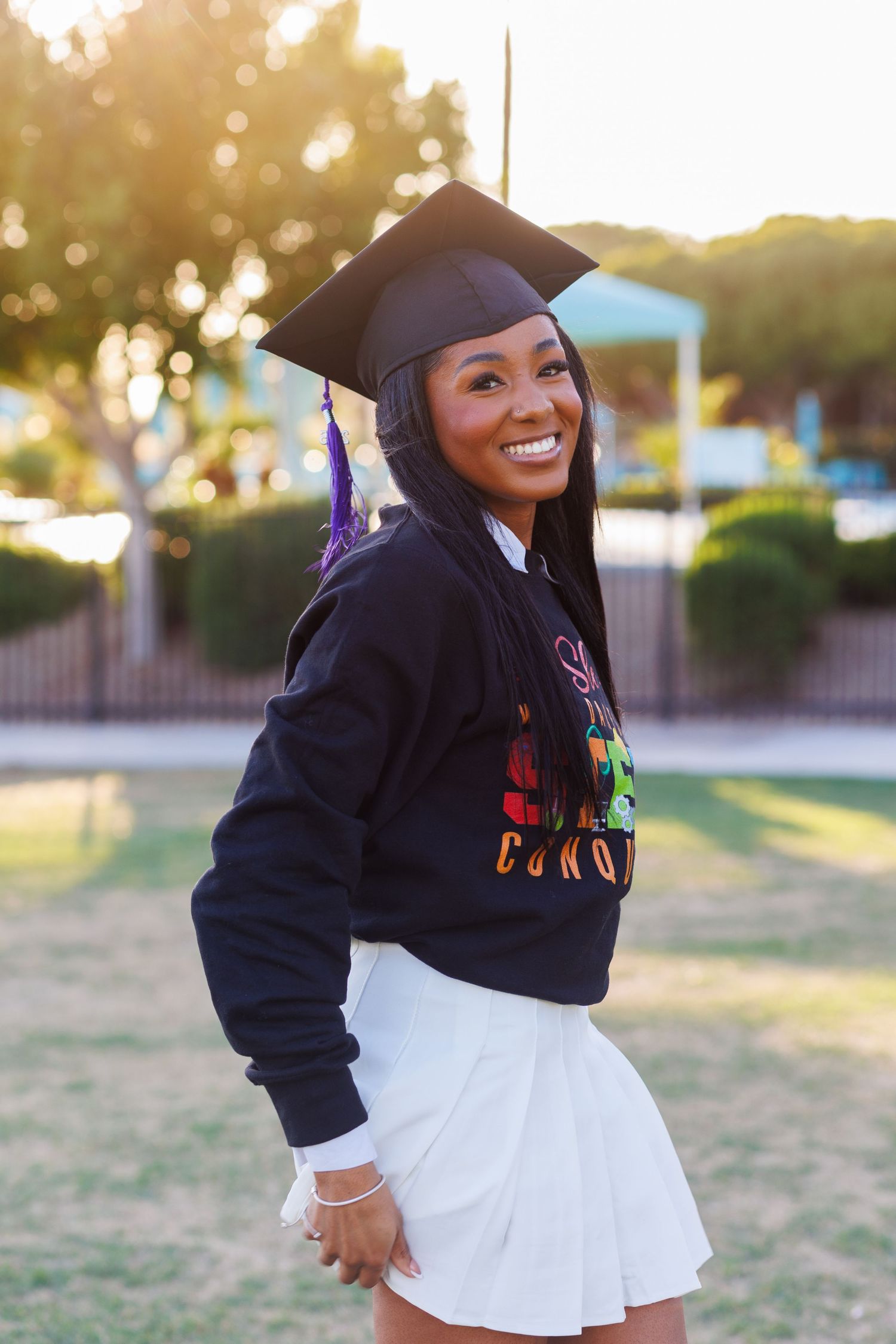 Smiling graduate in black cap and Grand Canyon University sweatshirt with white skirt at sunset.