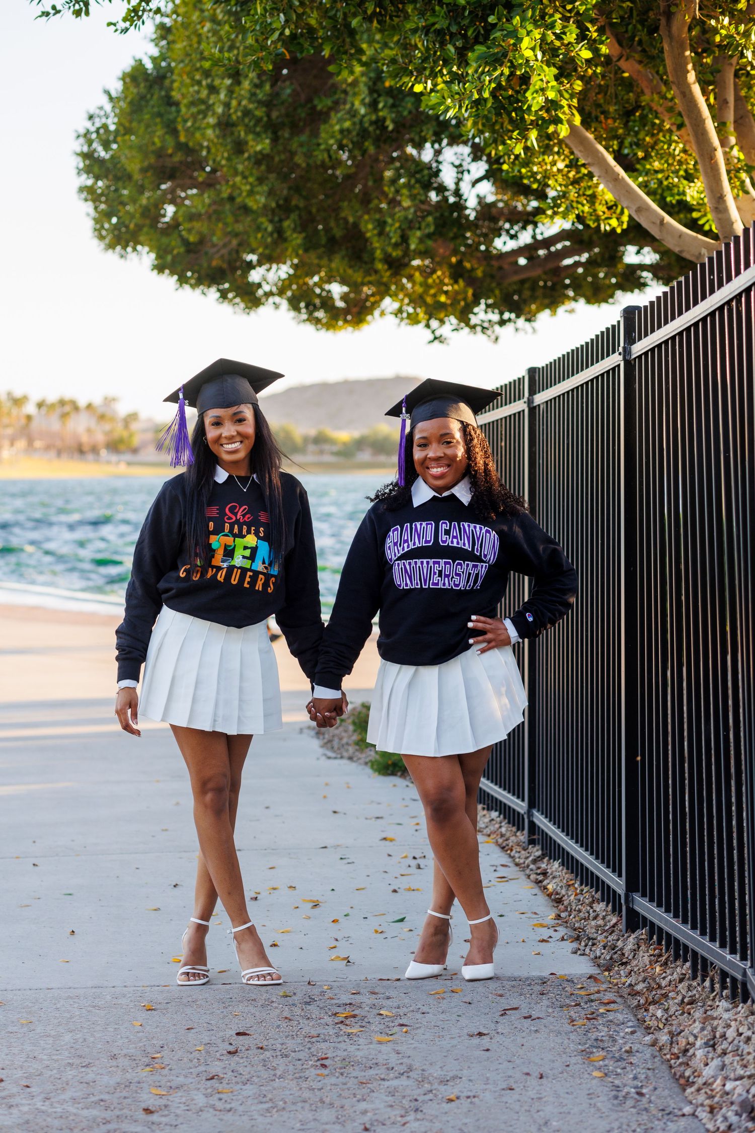 Two smiling graduates in black caps and Grand Canyon University sweatshirts hold hands outdoors.