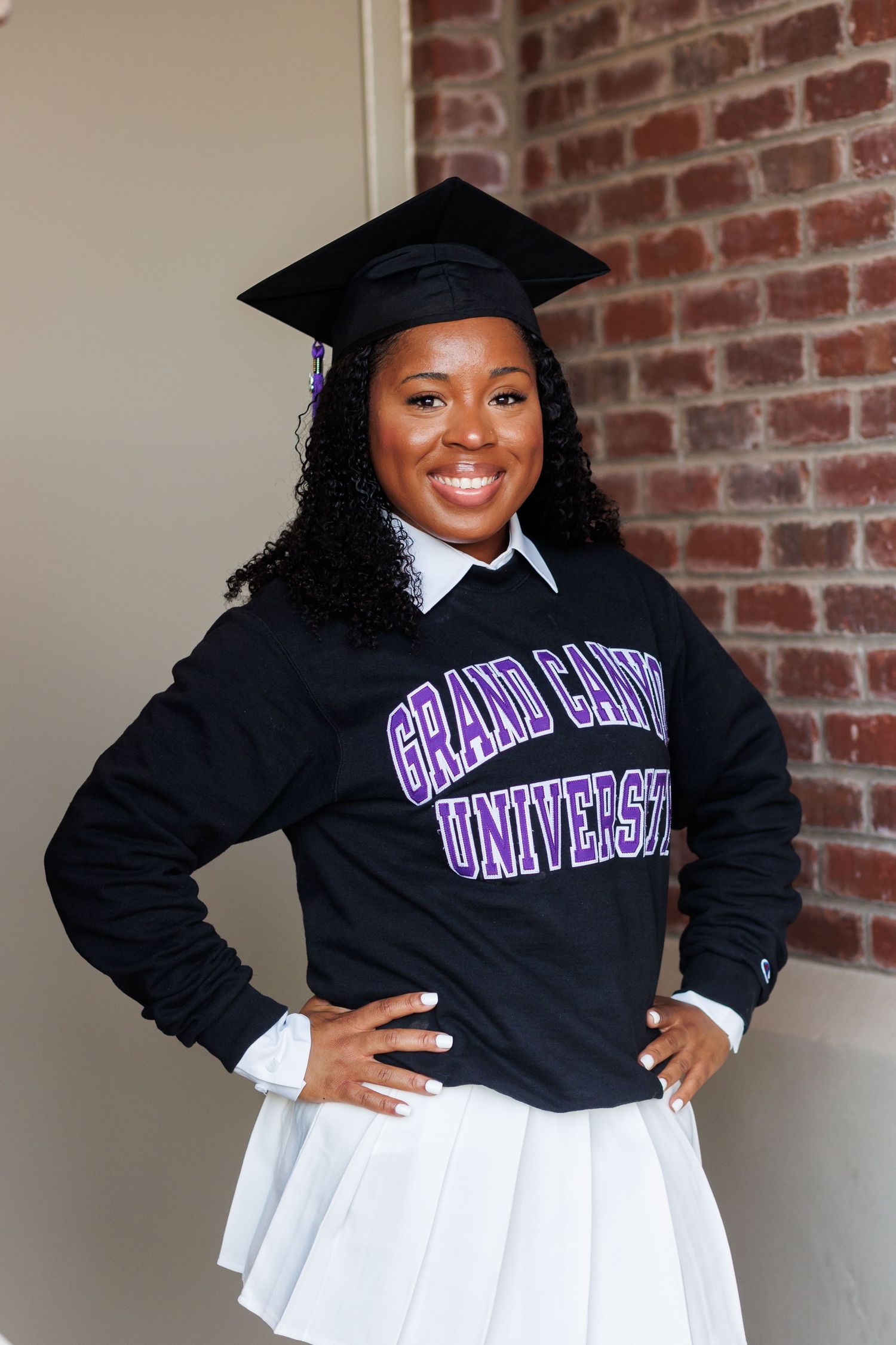 Smiling graduate poses in black Grand Canyon University sweatshirt, cap, and white skirt by brick wall.