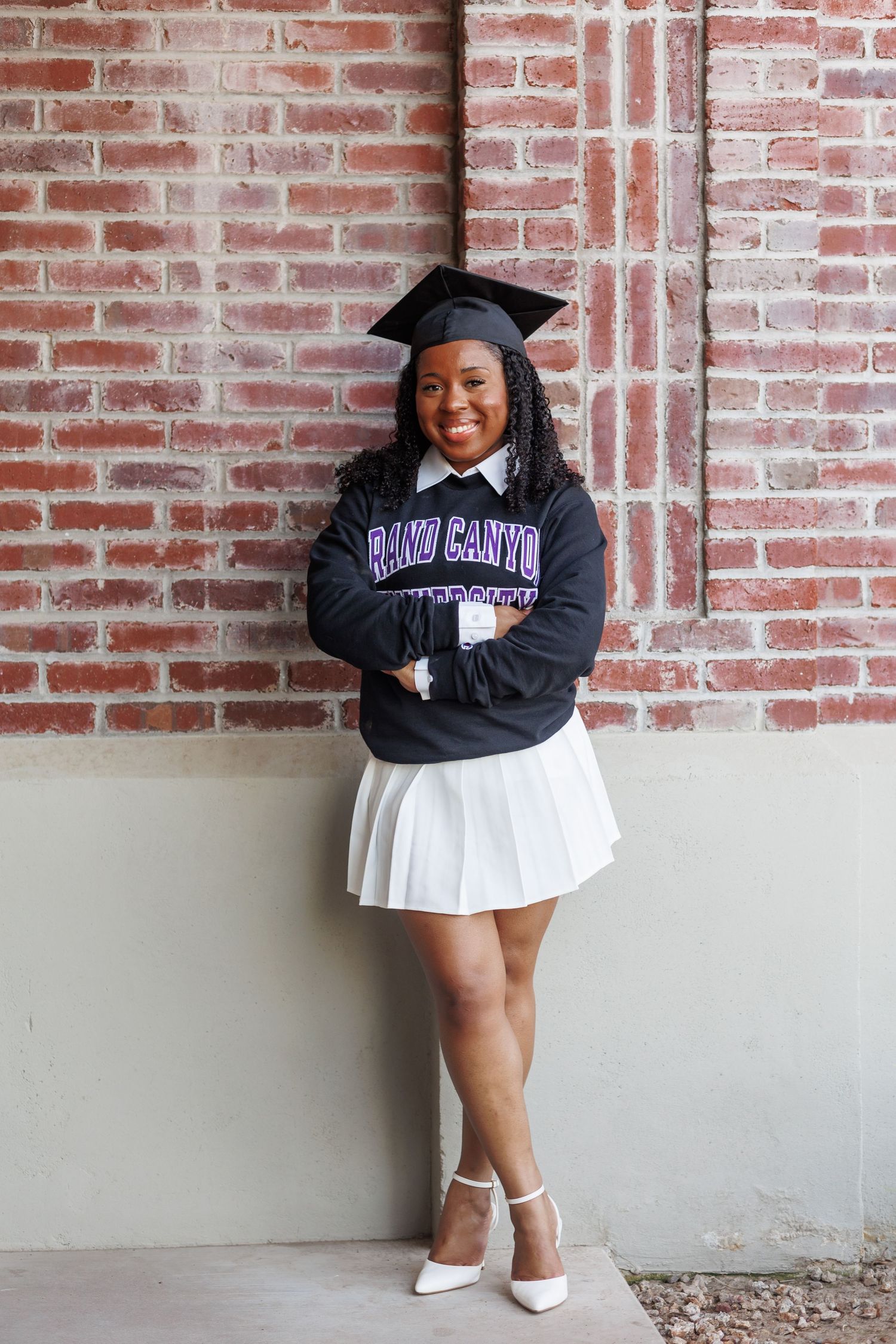 Graduate in Grand Canyon University sweatshirt and cap stands with arms crossed by red brick wall.