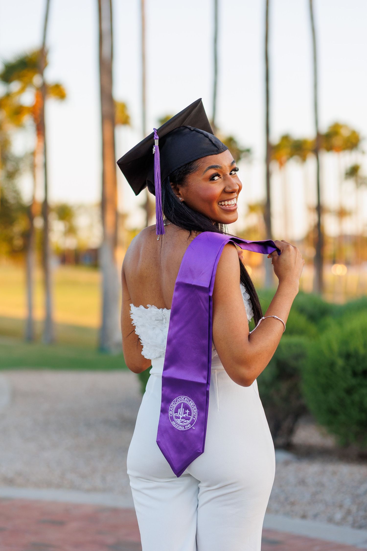 Smiling graduate in white outfit with purple sash and black cap poses outdoors near palm trees.