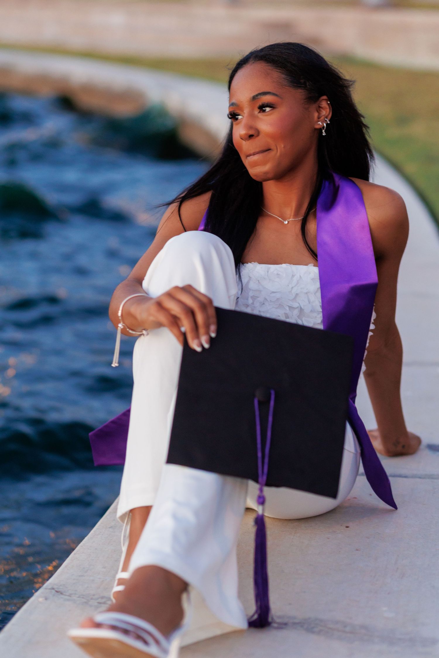 Young woman in white outfit with purple graduation sash sits by water holding black cap.